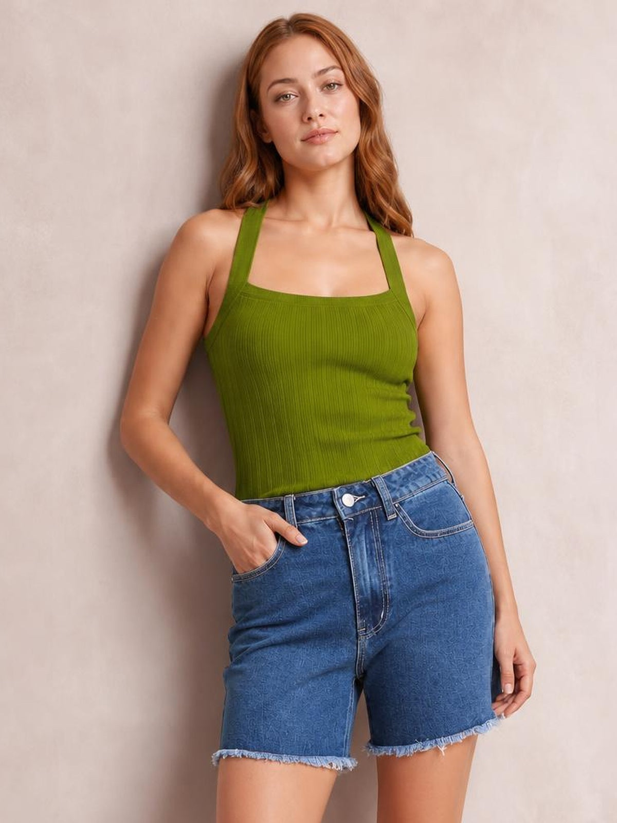 A woman wearing a ribbed olive green tank top and blue denim shorts, posing against a neutral beige wall.