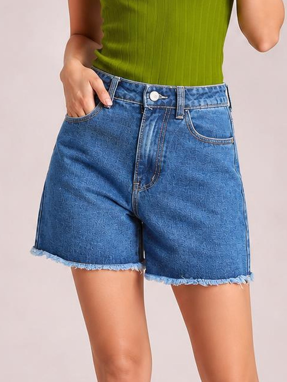 A model wears blue high-waist denim shorts with a button closure, front pockets, relaxed silhouette, and frayed raw hems, posing with one hand in a pocket.