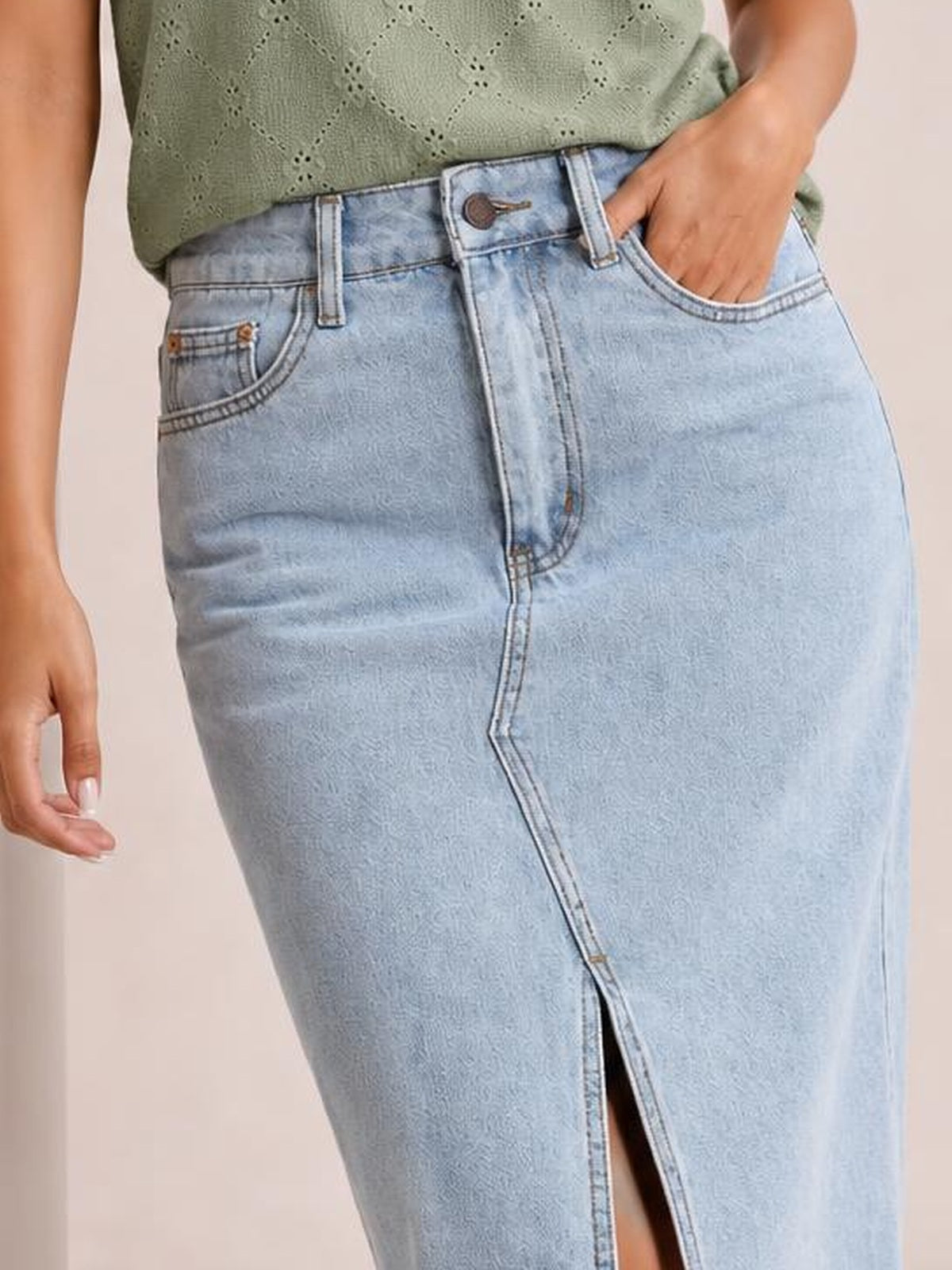 A model wears a light blue denim skirt with a fitted silhouette, front button closure, belt loops, functional pockets, and a center front slit.