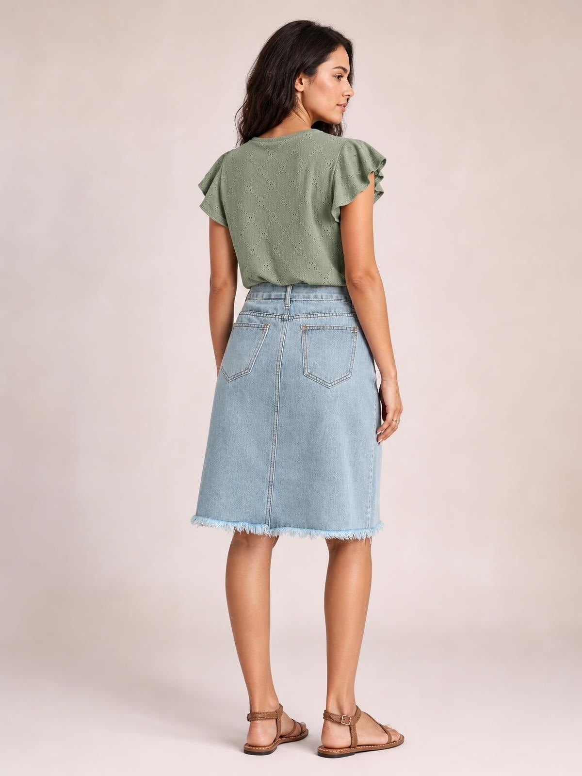 A model stands turned away in a light blue denim skirt with a straight midi silhouette, back patch pockets, center seam, and frayed raw hem.