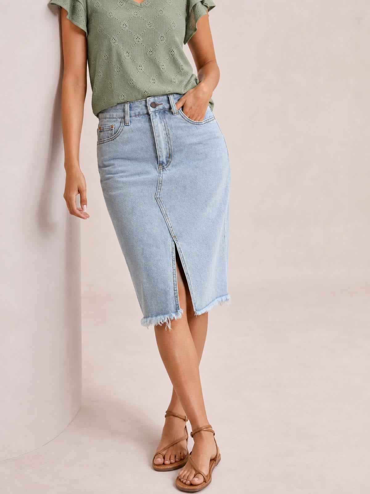 Model wears a light-wash denim skirt with a buttoned high waist, pockets, front slit, fitted midi silhouette, and raw frayed hem.