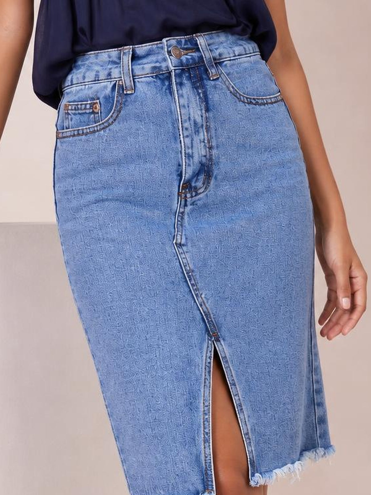 Model wearing a light blue raw hem slit denim skirt with a fitted silhouette, front button closure, belt loops, curved pockets, and center-front slit.