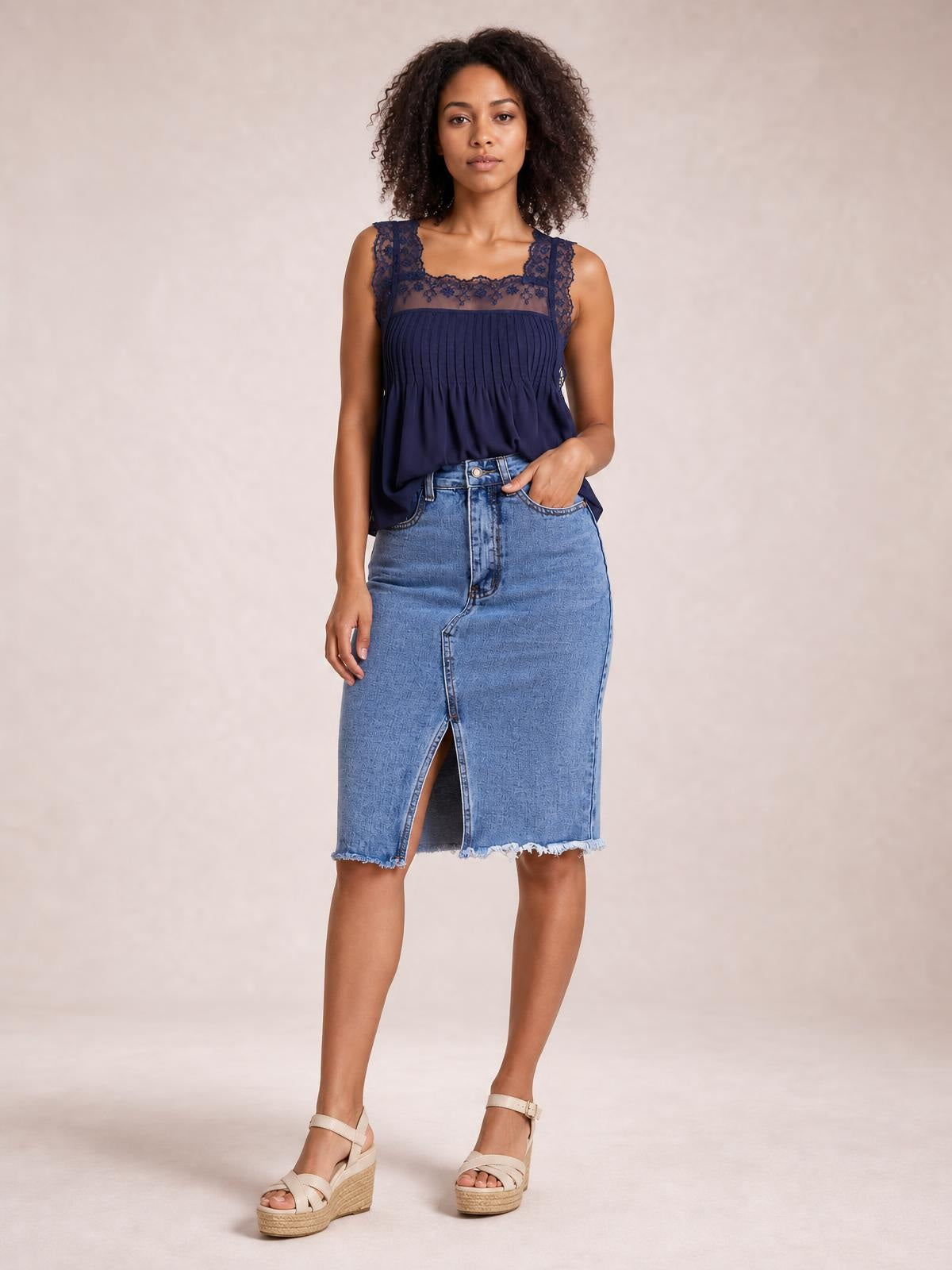 A model stands in a light blue raw hem denim skirt with a straight midi silhouette, front button closure, pockets, and a center slit, one hand in a pocket.