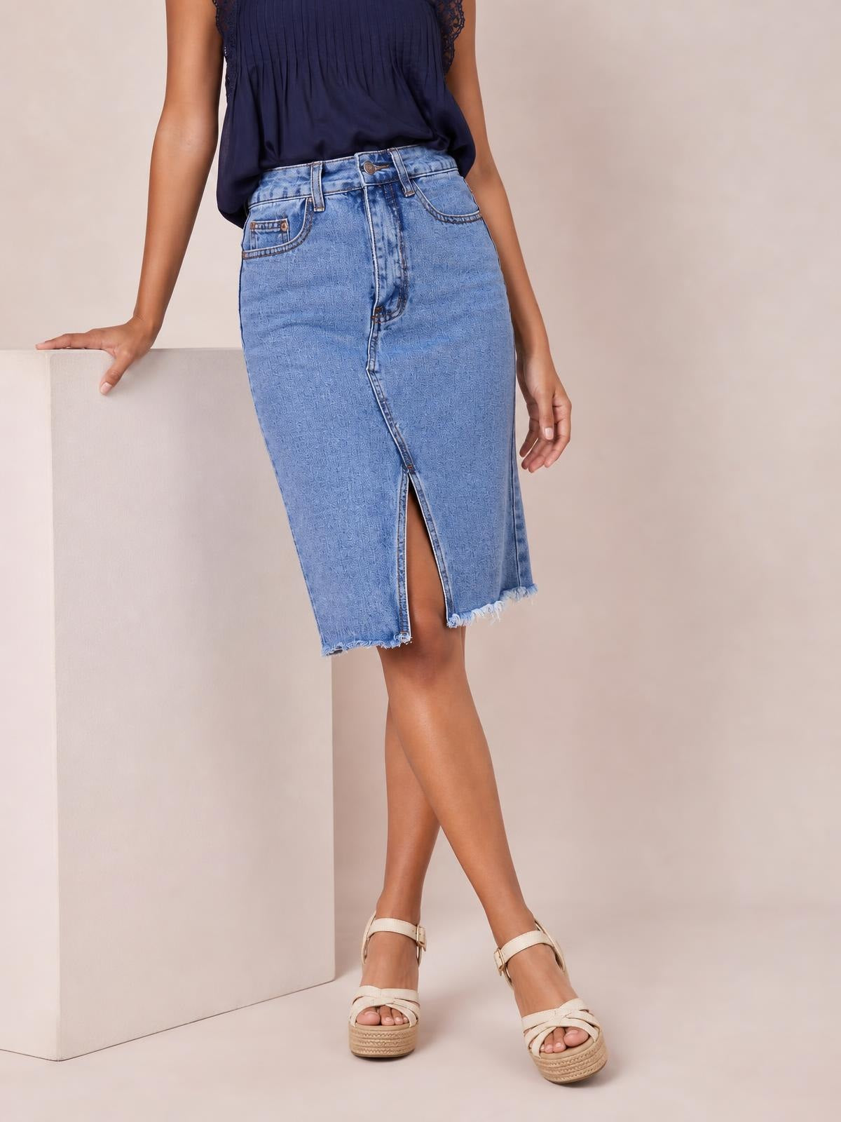 Model poses with one hand on a pedestal, wearing a light-wash denim midi skirt with a fitted silhouette, button closure, front slit, pockets, and raw hem.