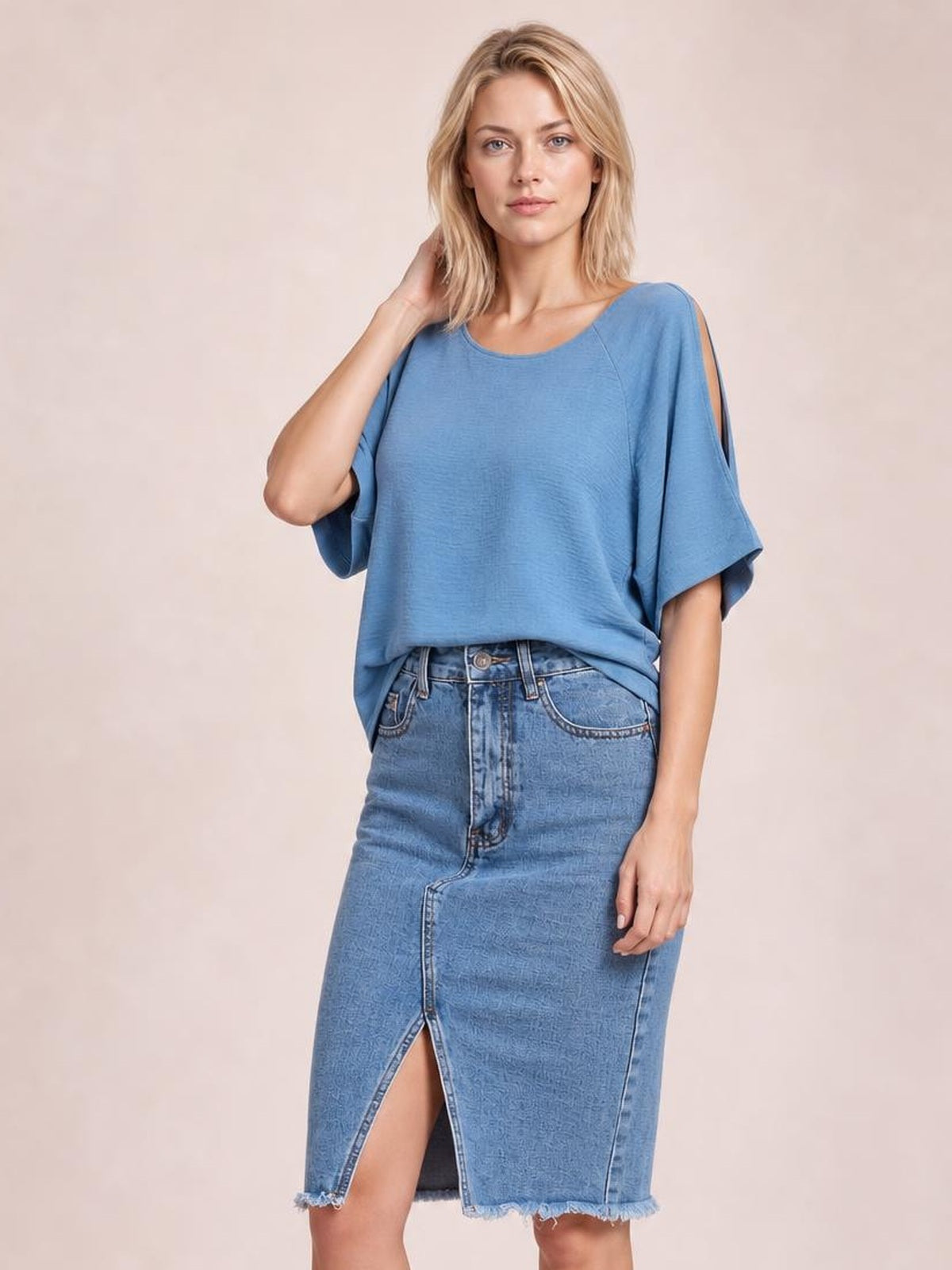 Model poses in a light blue denim skirt with a buttoned front, pockets, straight silhouette, front slit, and raw frayed hem.