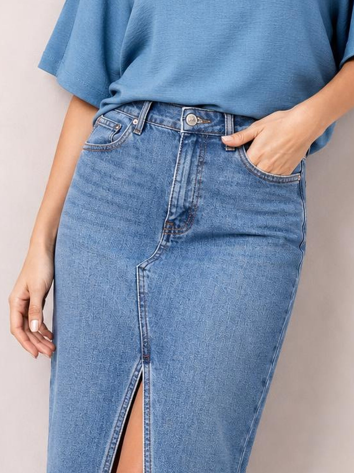 Model wearing a light blue raw hem slit denim skirt with a fitted silhouette, button closure, front pockets, and a front slit.