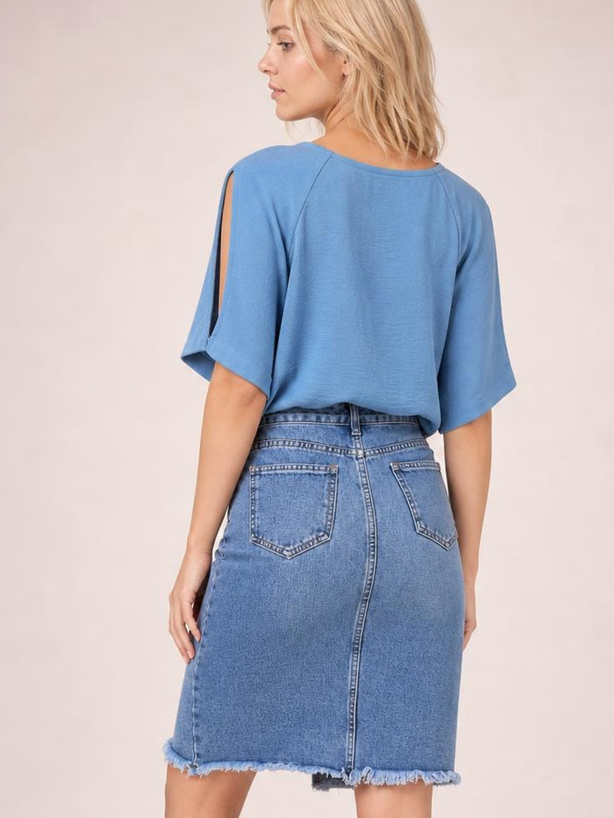 A model stands facing away in a blue raw hem denim skirt with a straight midi silhouette, back patch pockets, and a center-back seam.