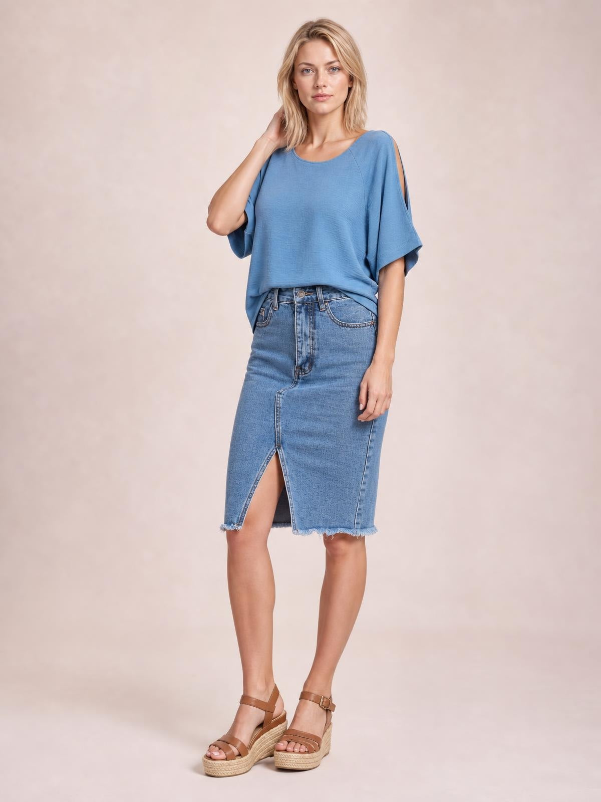 A model poses in a light blue raw hem slit denim skirt with a fitted midi silhouette, front button closure, and visible pockets.