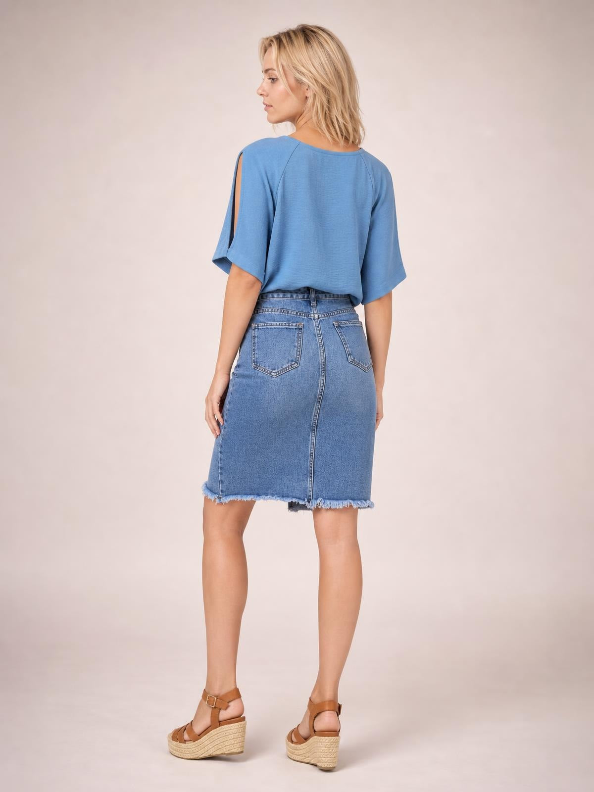 A model poses turned away in a blue denim skirt with a fitted knee-length silhouette, back pockets, center seam, and edgy raw frayed hem.