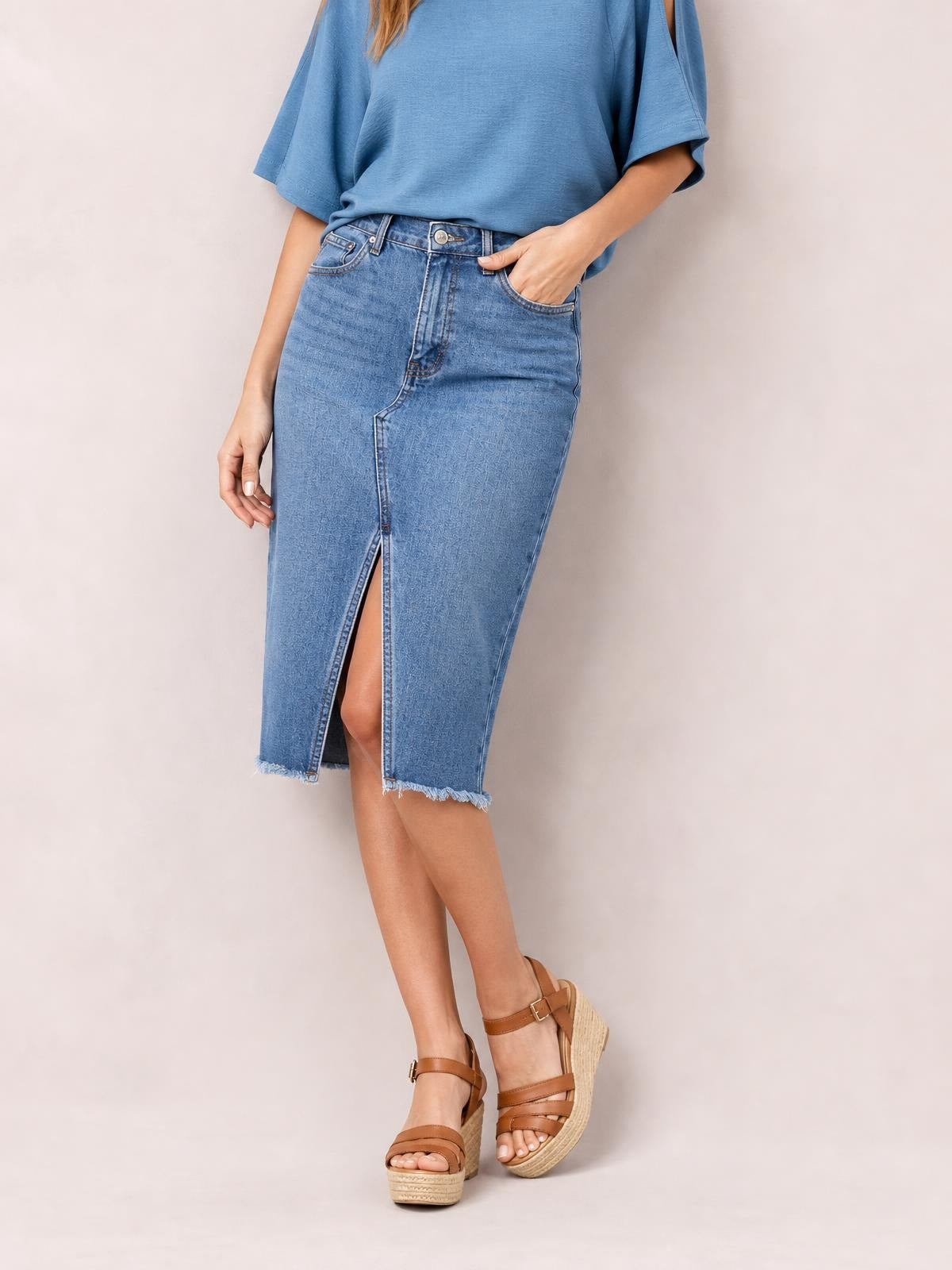 Model wears a light blue raw hem slit denim skirt with a fitted midi silhouette, button closure, front pockets, and a front slit.