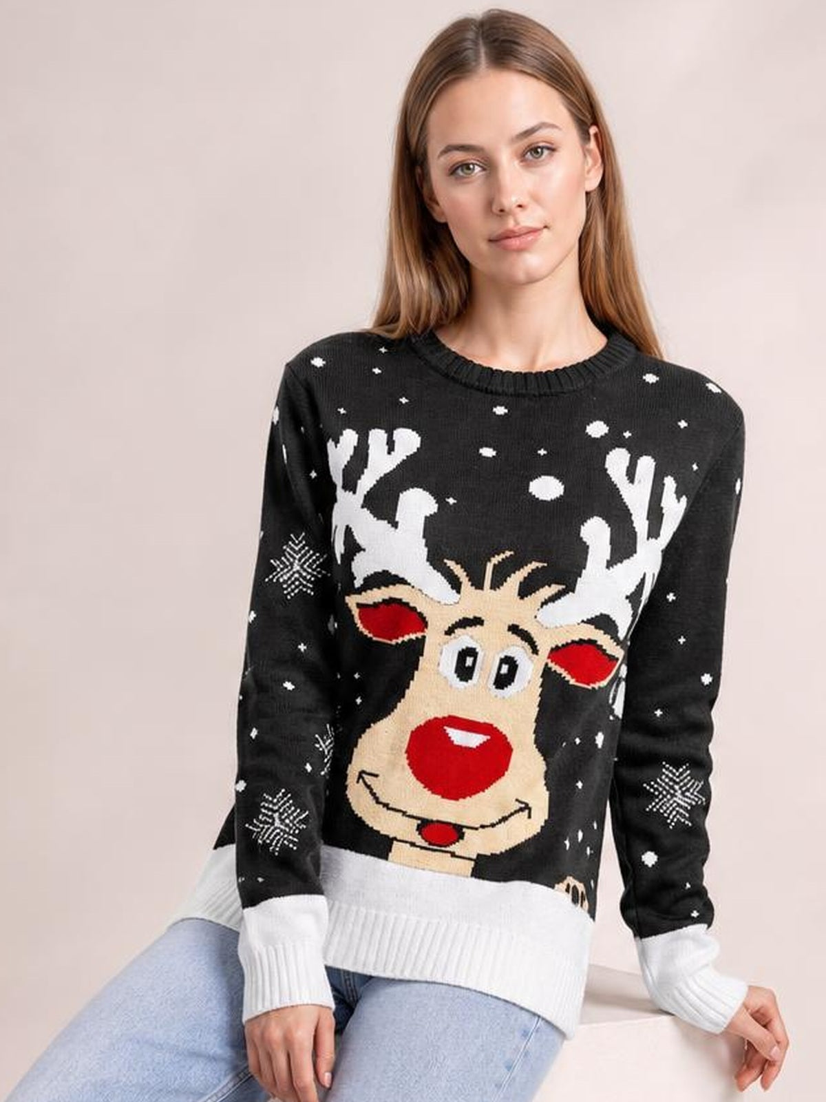 Seated model wears a black reindeer round neck sweater with a festive knit motif, white snowflakes, ribbed cuffs and hem, and a relaxed silhouette.