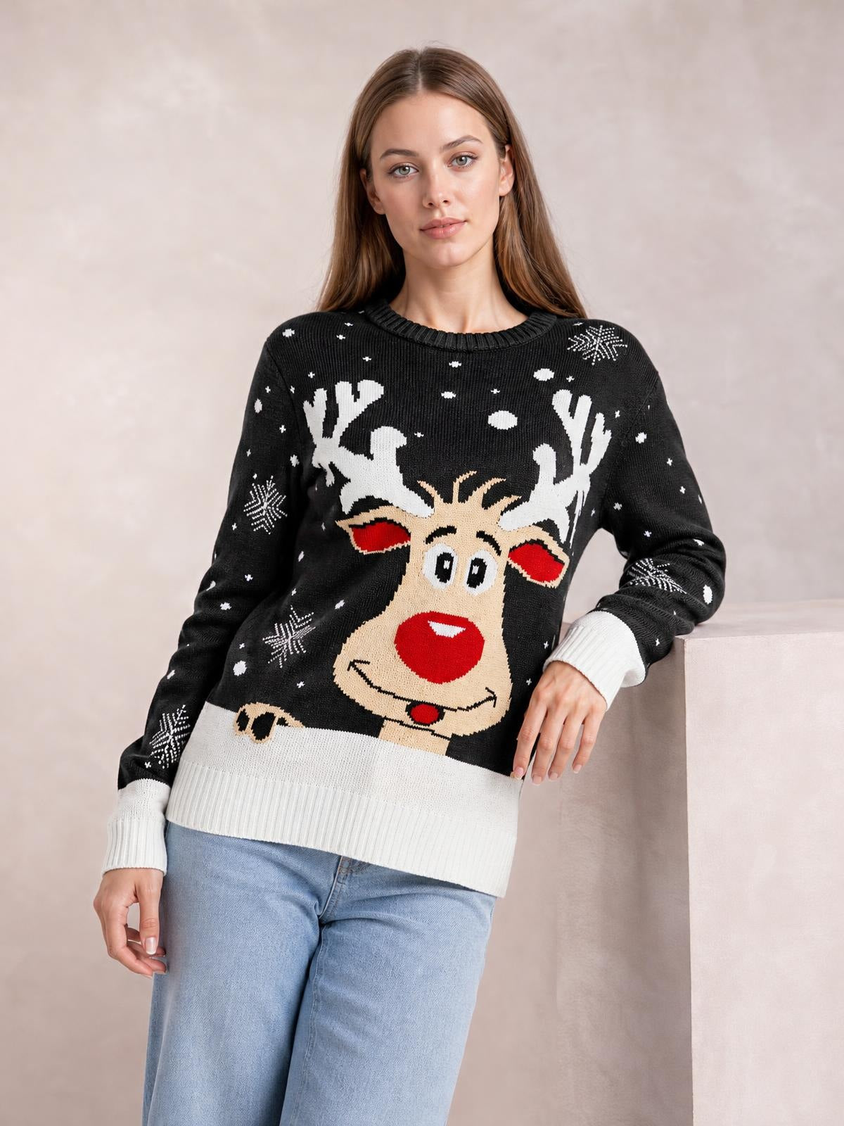 Model leans against a ledge wearing a black Reindeer round neck sweater with a festive reindeer graphic, white ribbed cuffs and hem, and a relaxed silhouette.
