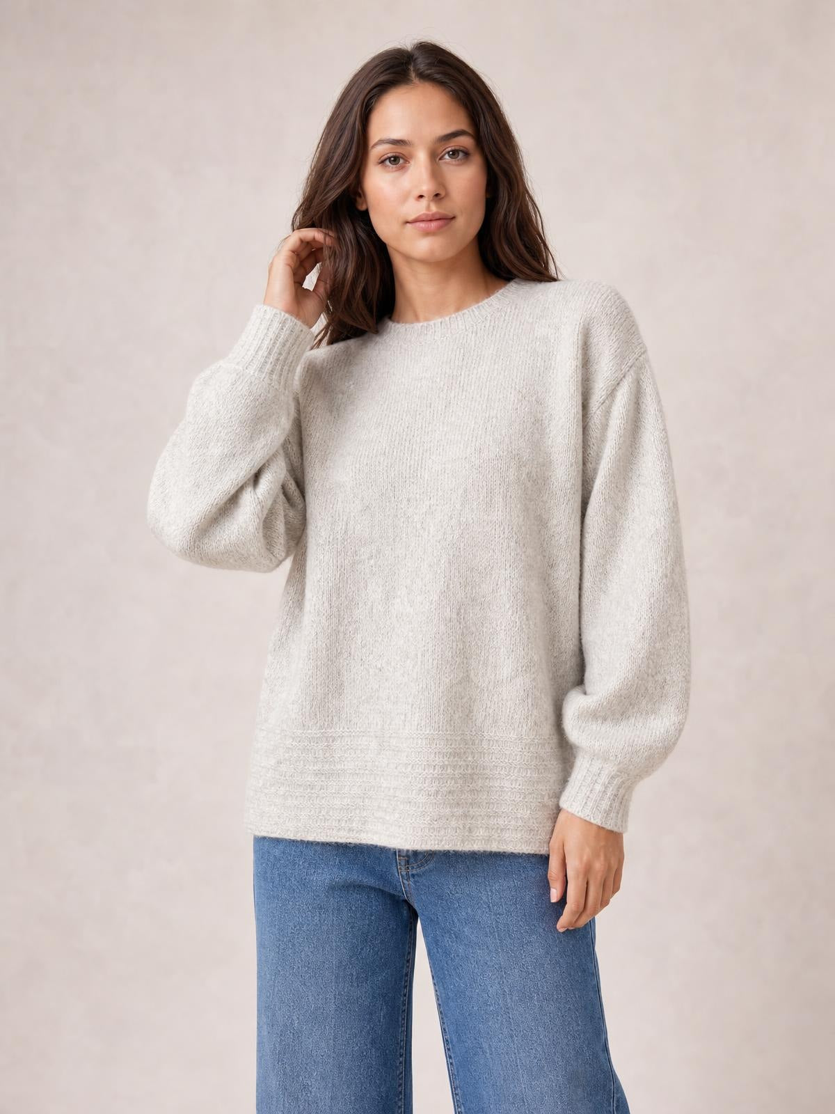 Model wearing a light beige relaxed fit round neck sweater with dropped shoulders, long sleeves, ribbed cuffs and hem, posing with one hand near her hair.