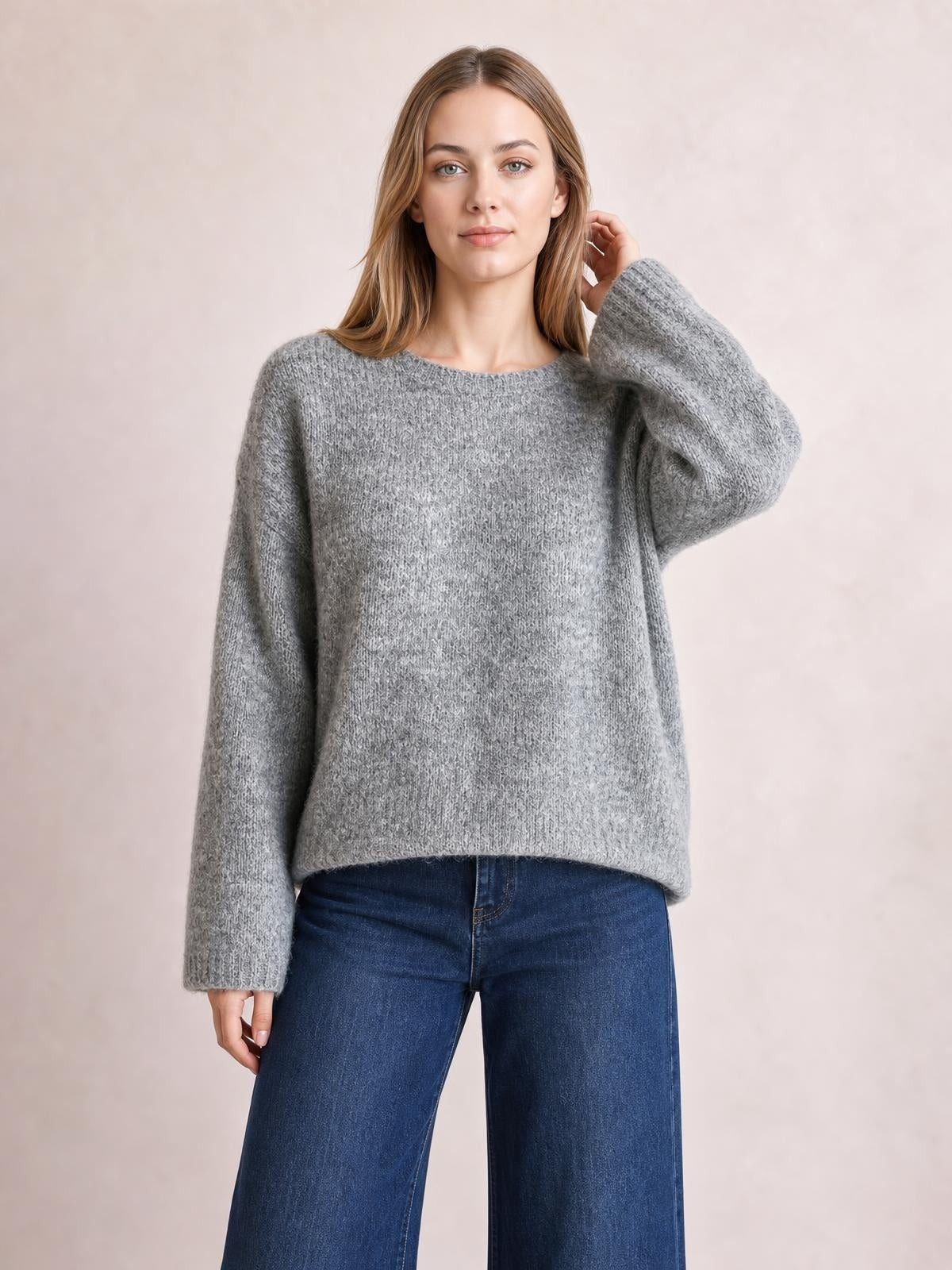 Model wearing a gray relaxed fit round neck sweater with a soft textured knit, long sleeves, and a loose straight silhouette, touching her hair.