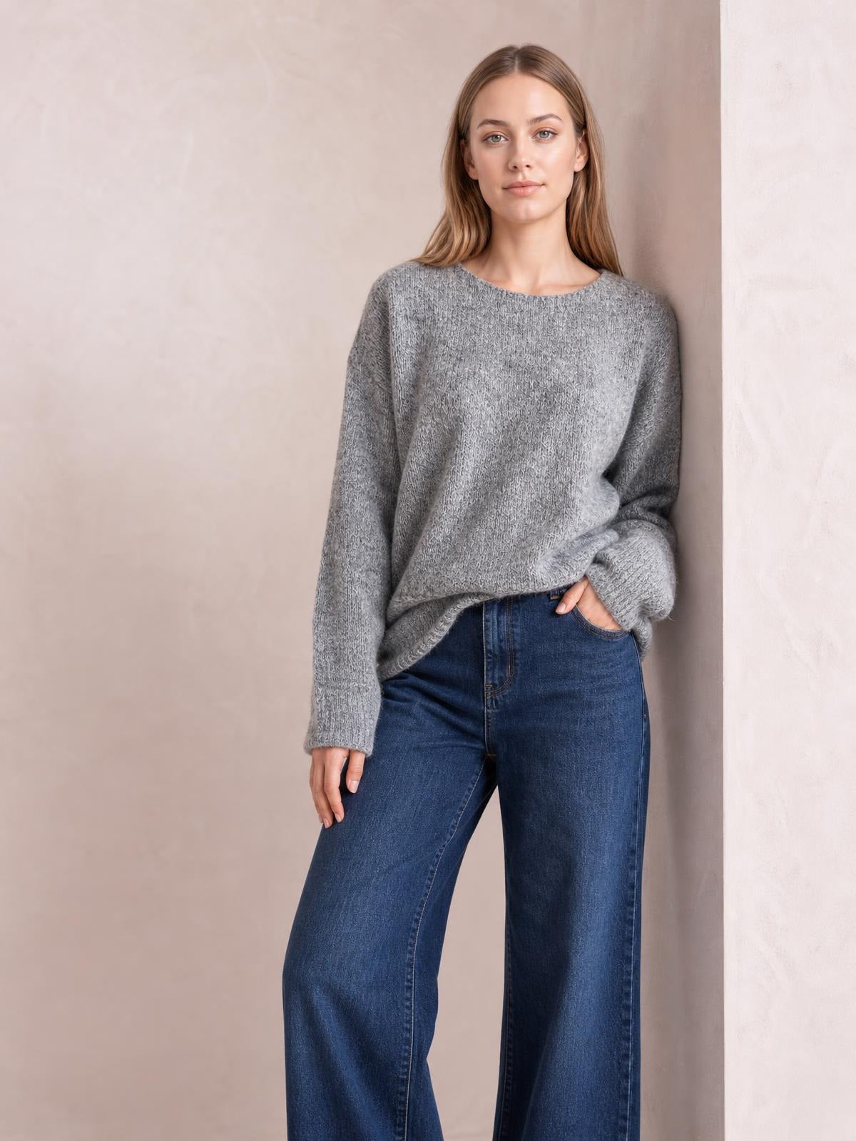 Model wearing a relaxed fit gray round neck sweater with long sleeves and ribbed cuffs, standing with one hand in her jeans pocket.