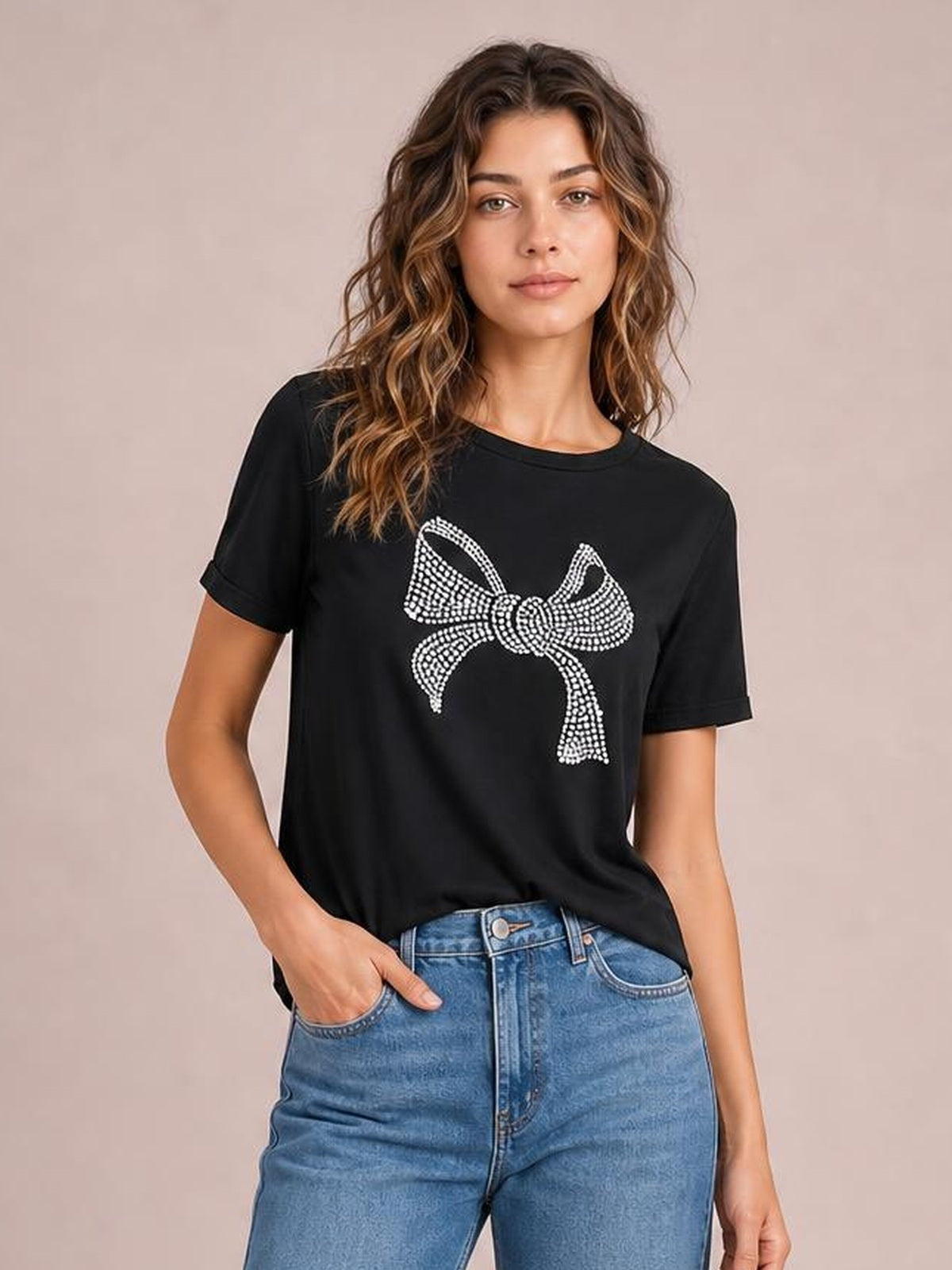Model wearing a black round-neck short-sleeve T-shirt with a large rhinestone bow graphic, relaxed straight silhouette, and one hand in her jeans pocket.