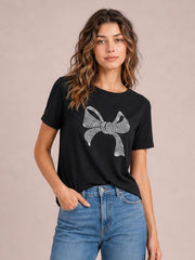 Model wearing a black round-neck short-sleeve T-shirt with a large rhinestone bow graphic, relaxed straight silhouette, and one hand in her jeans pocket.