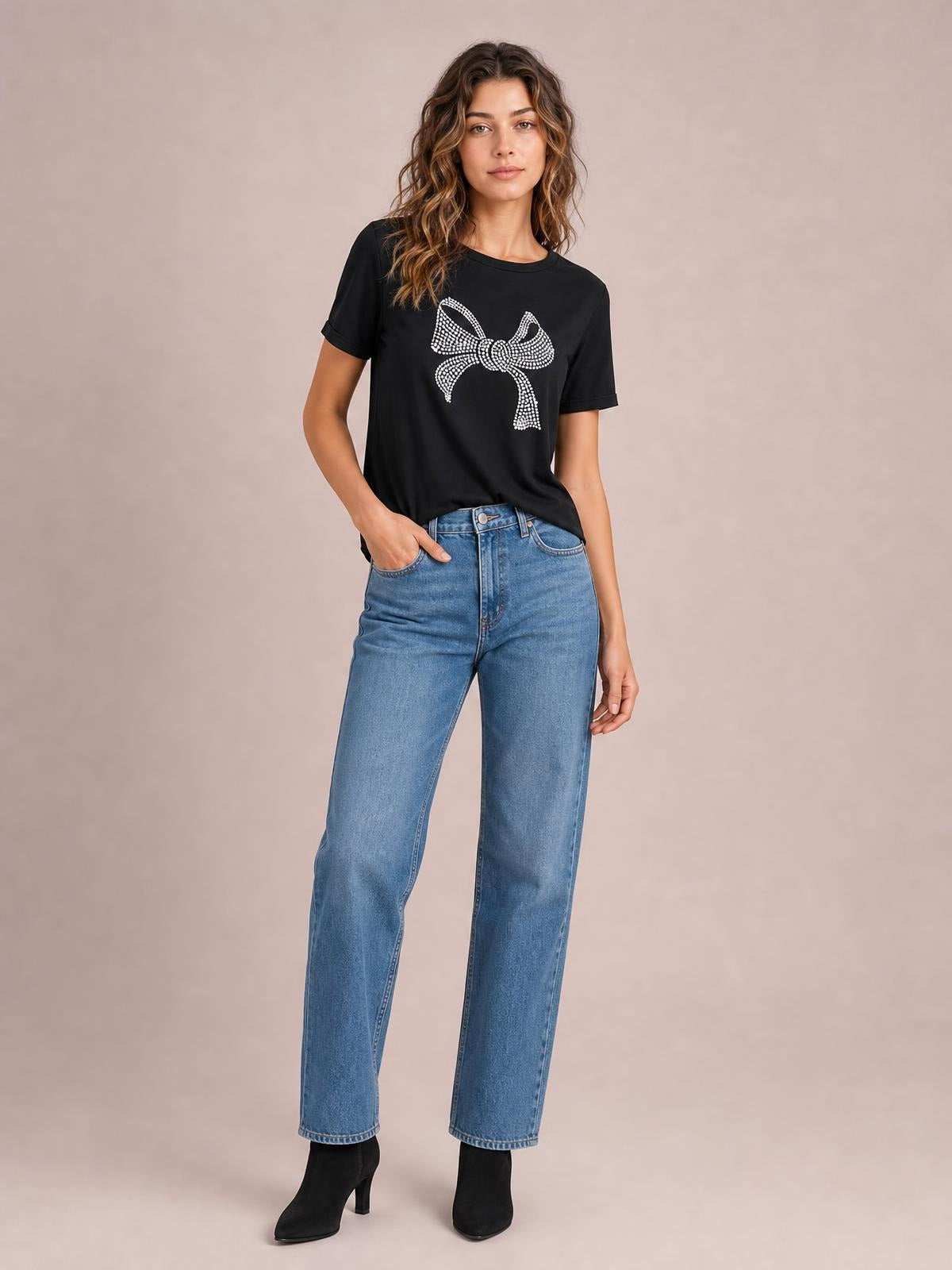 A model wears a black round-neck short-sleeve T-shirt with a sparkling rhinestone bow graphic and relaxed silhouette, posing with one hand in her jeans pocket.