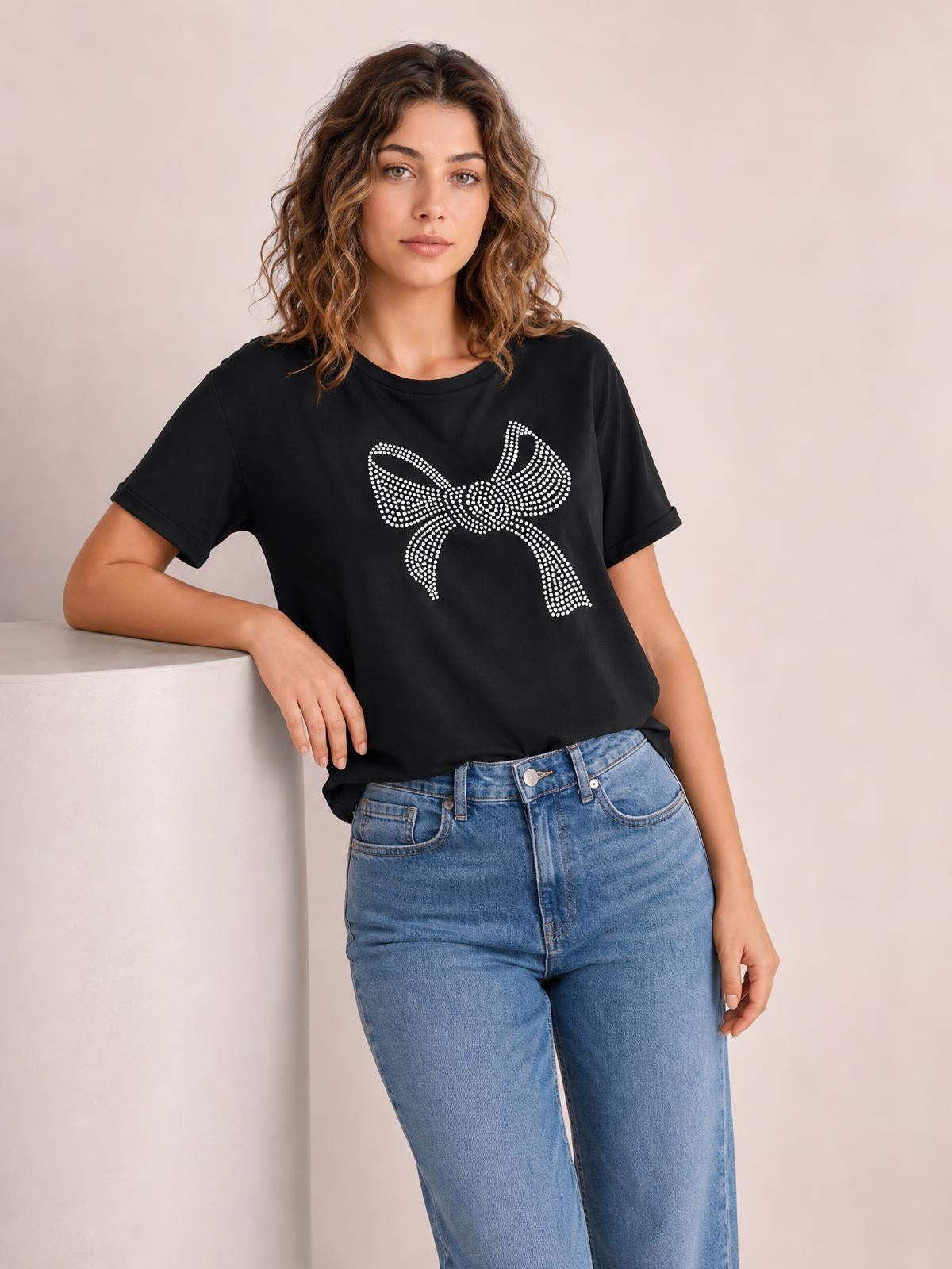 Model leans against a ledge wearing a black round-neck short-sleeve T-shirt with a sparkling rhinestone bow graphic and relaxed silhouette.