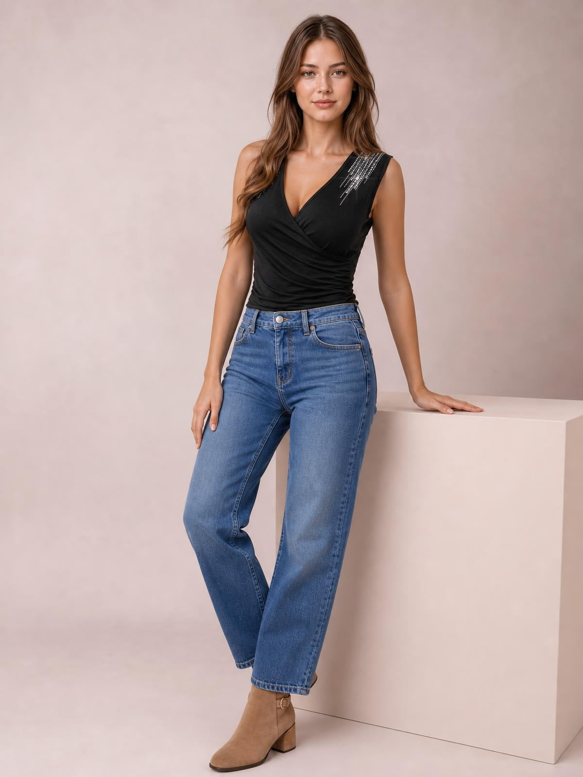 A model stands in a fitted black surplice tank with sleeveless styling, ruched construction, and rhinestone shoulder detail, paired with jeans.