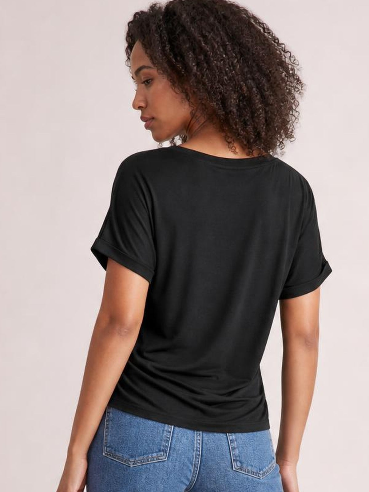 Model turned slightly away to show the back of a black short-sleeve T-shirt with rolled cuffs, round neckline, and relaxed straight silhouette.