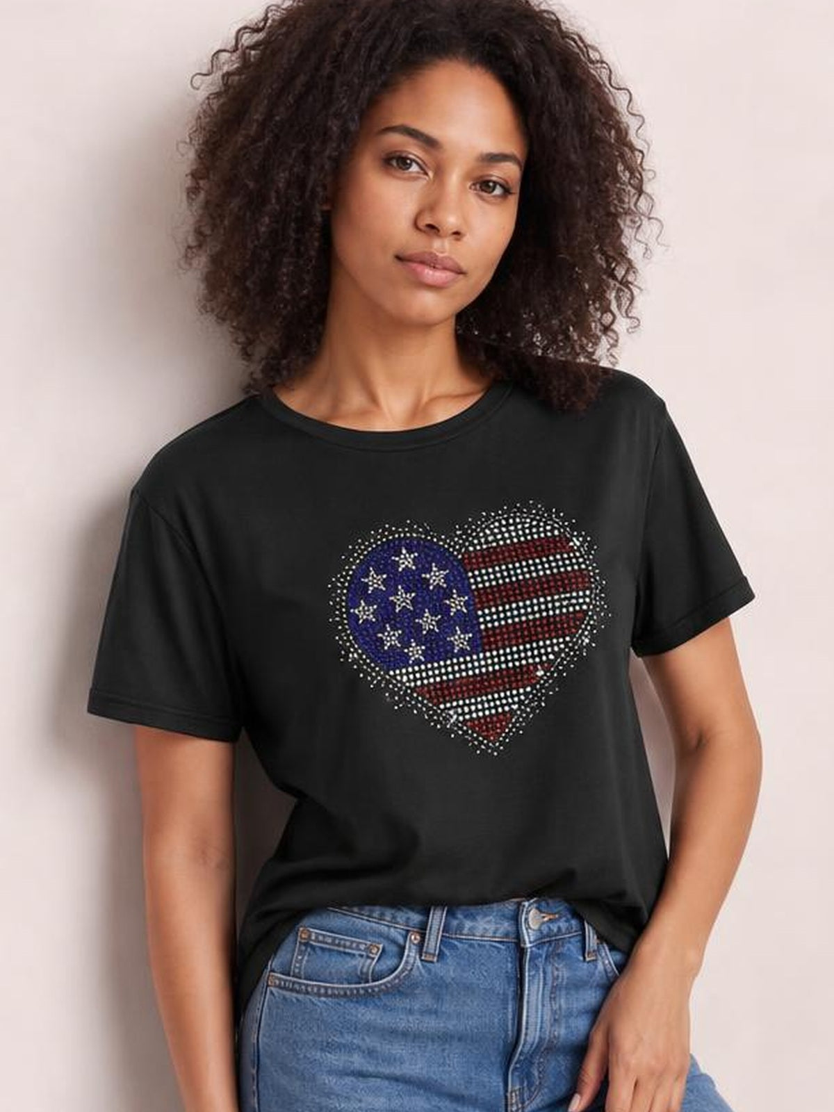 A model poses in a black round-neck short-sleeve T-shirt with a relaxed silhouette and sparkling rhinestone heart featuring an American flag design.