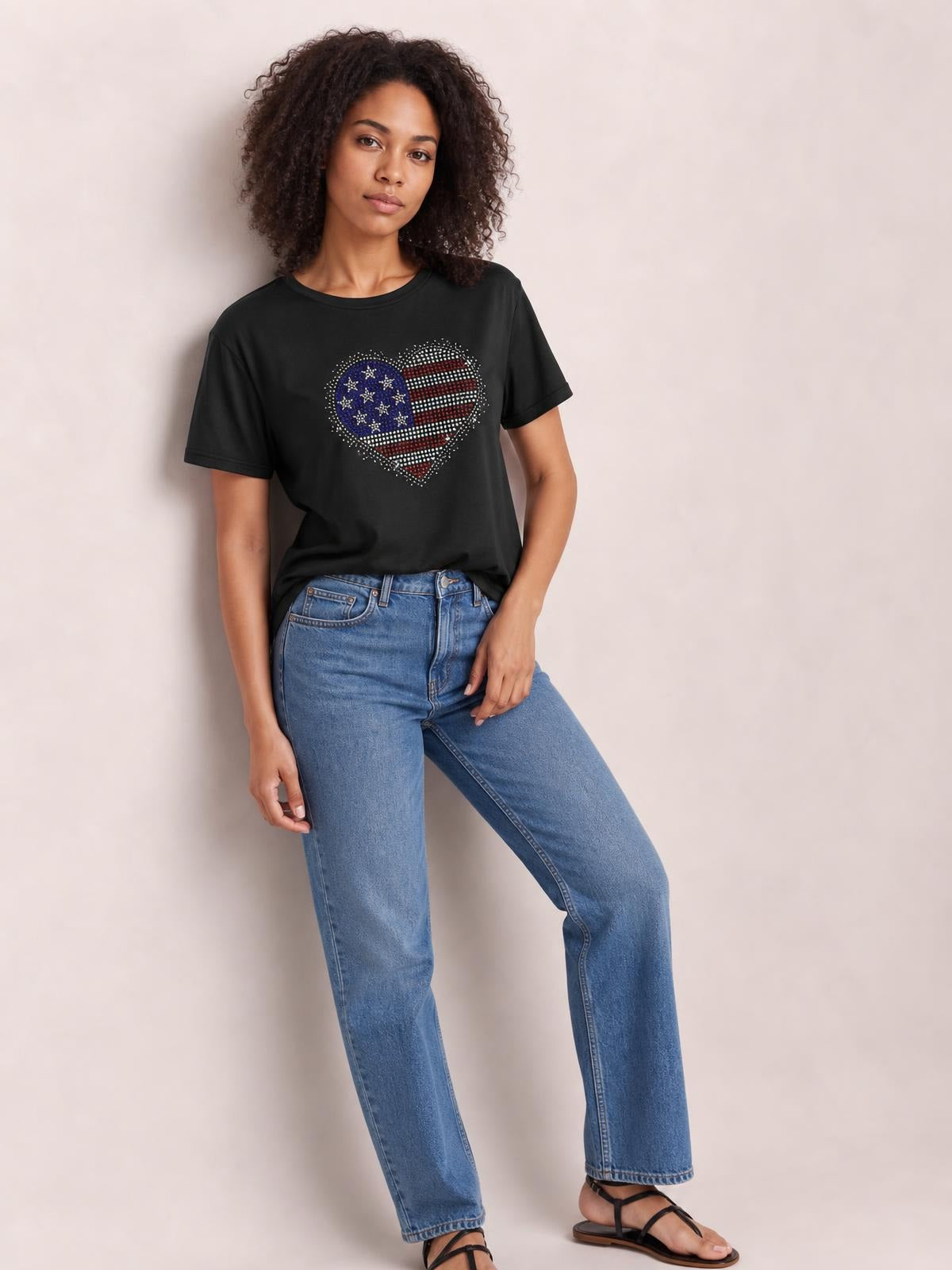 A model poses in a black round-neck short-sleeve T-shirt with a relaxed silhouette and sparkling rhinestone heart graphic, one leg bent.