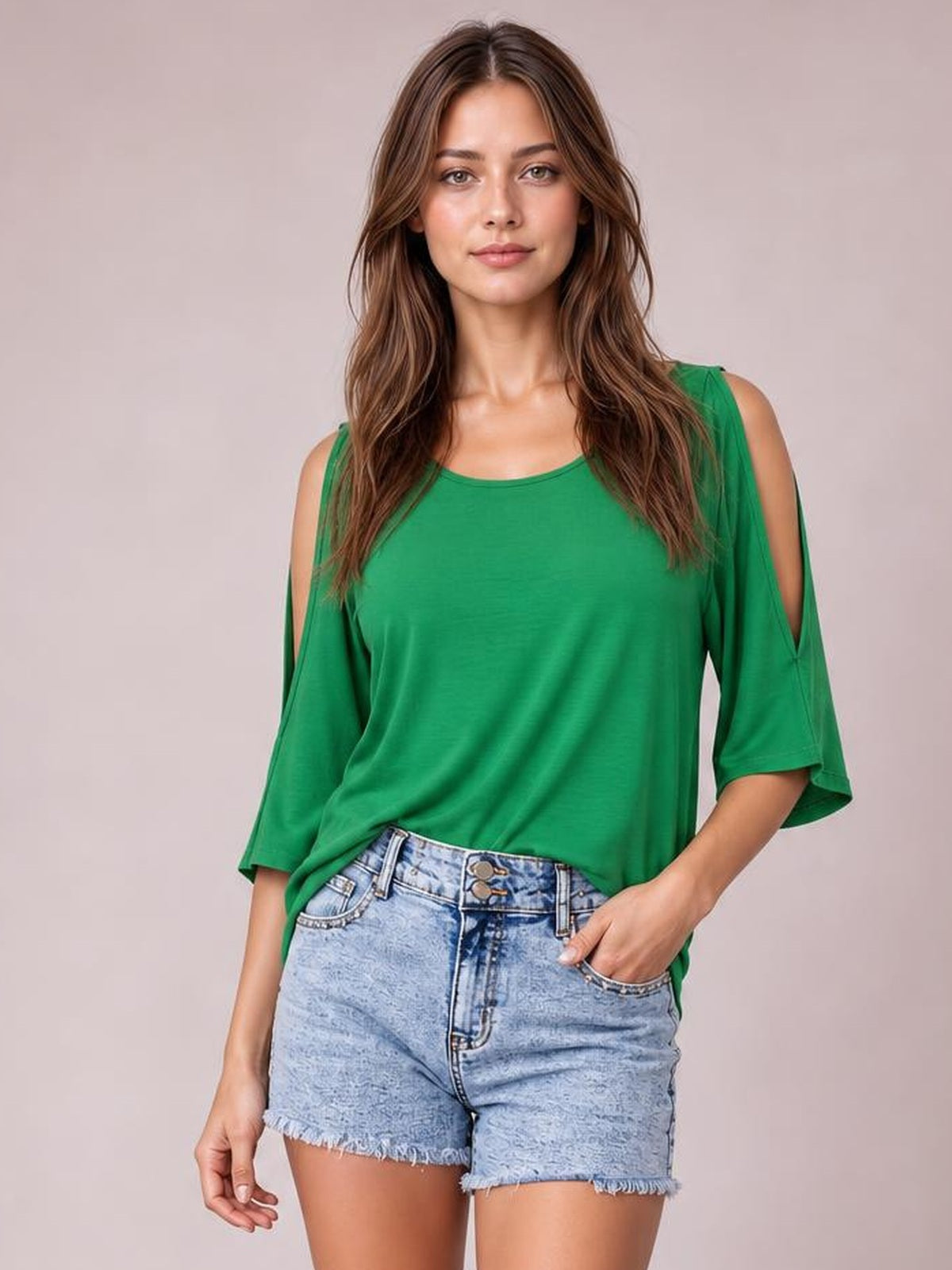 A woman modeling a bright green cold-shoulder top with three-quarter sleeves and light-wash denim shorts against a solid light beige studio background.
