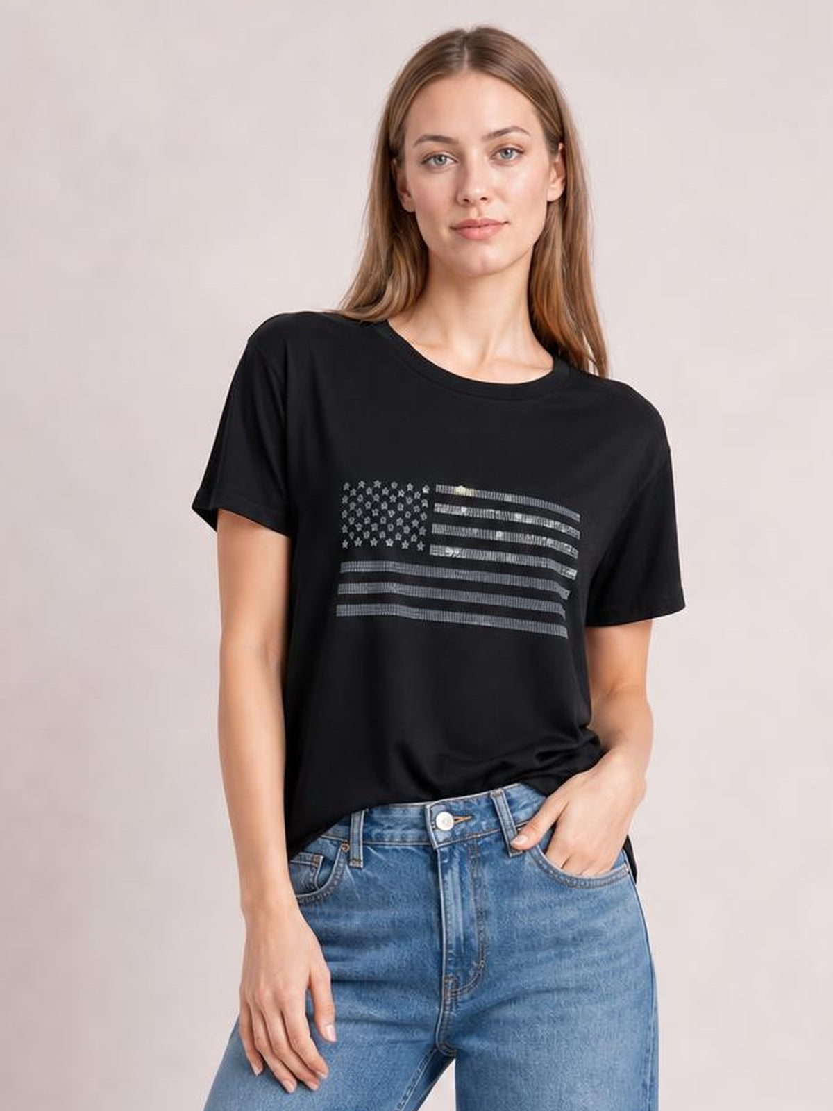 Model wearing a black round-neck short-sleeve T-shirt with a rhinestone US flag graphic, relaxed silhouette, and one hand resting in her jeans pocket.