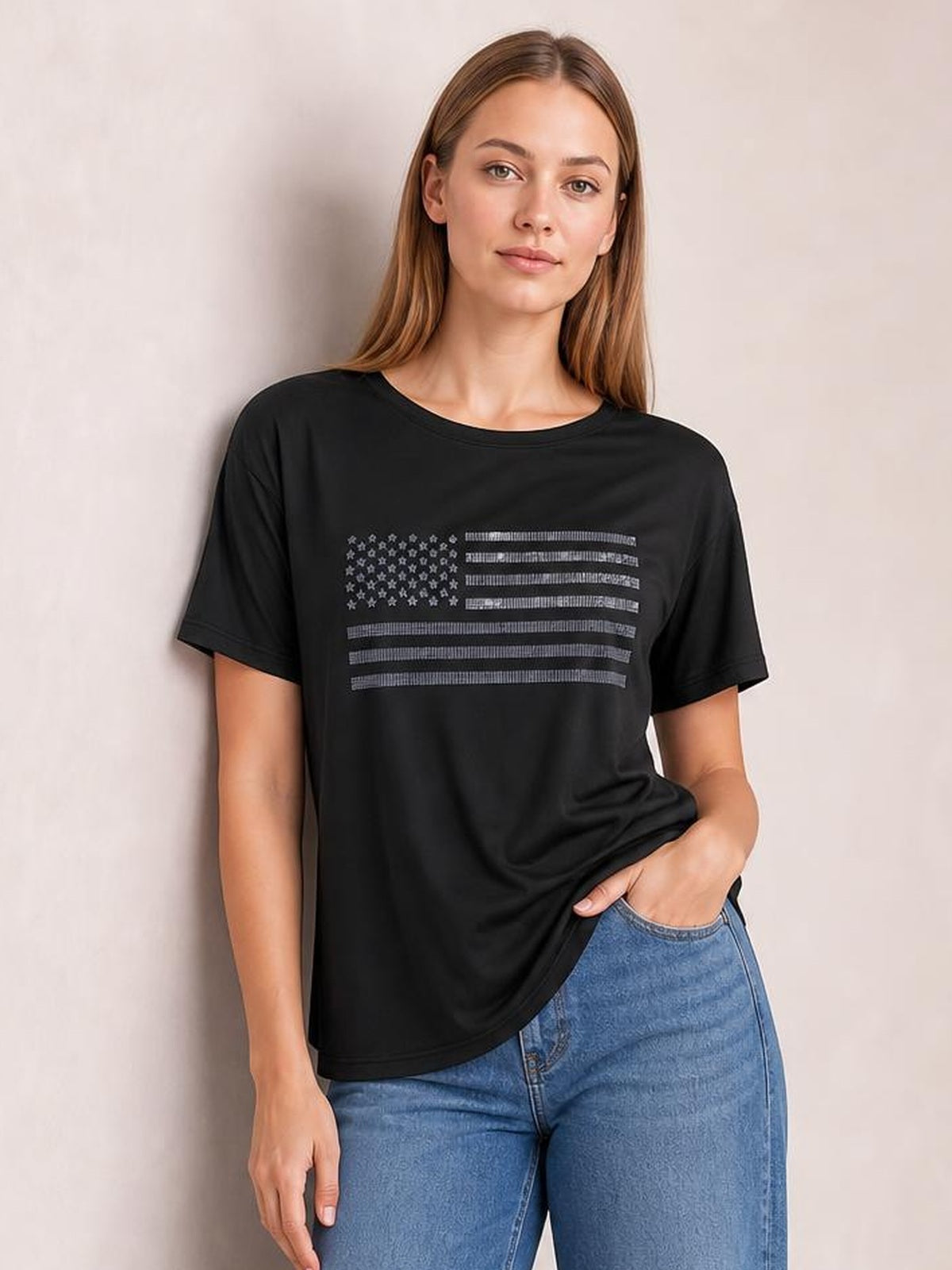 Model wears a black round-neck short-sleeve T-shirt with a sparkling rhinestone US flag graphic and relaxed silhouette, gently lifting the hem at one side.