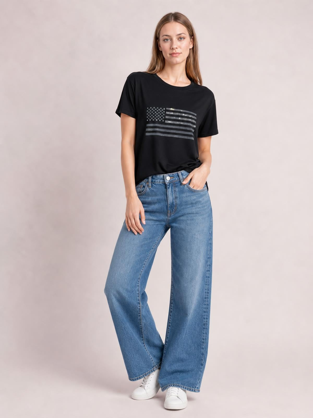 Model wears a black rhinestone US flag T-shirt with a round neck and short sleeves, posing casually with one hand in her jeans pocket.