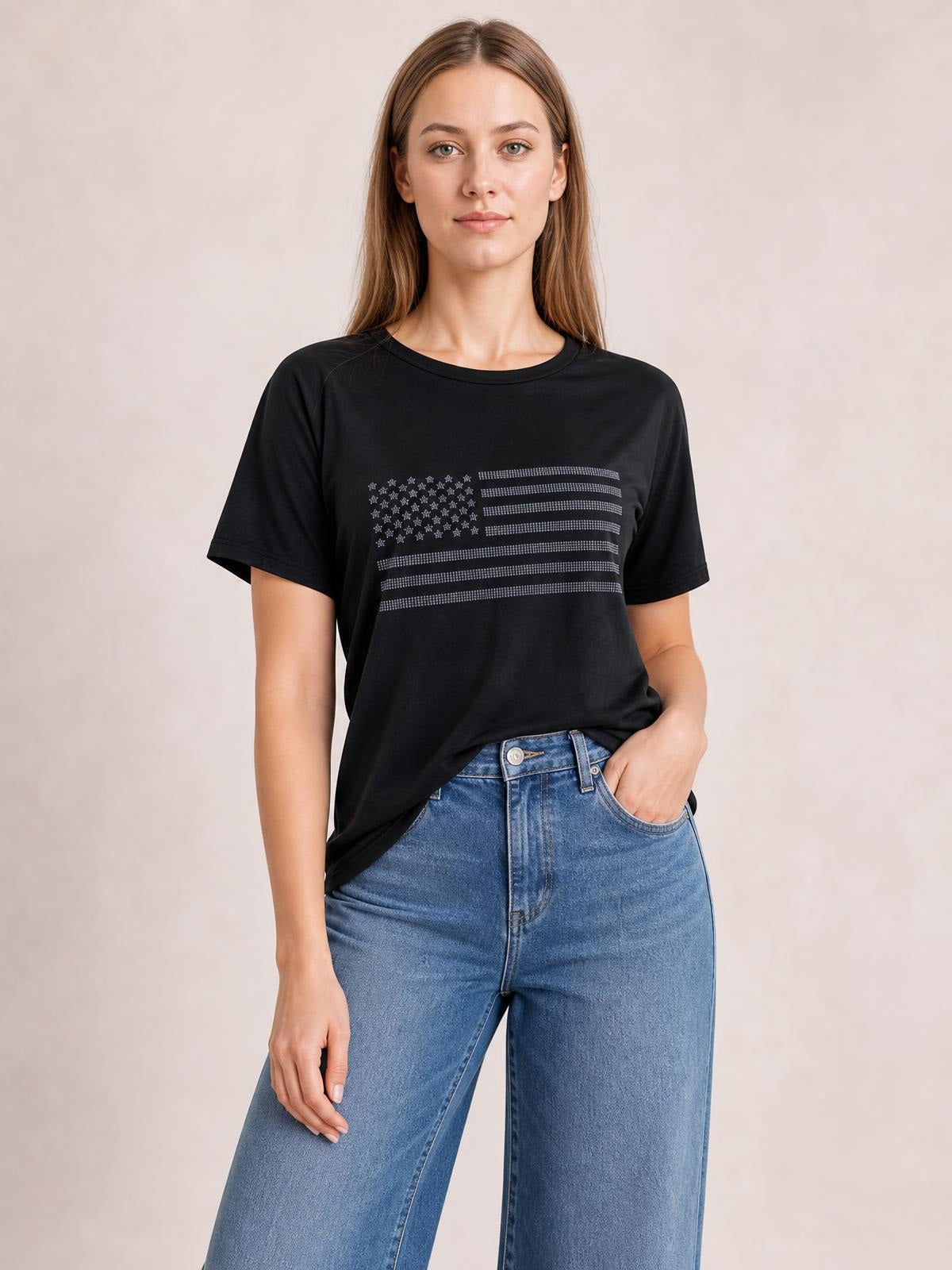 A woman stands in a black round-neck short-sleeve T-shirt with a rhinestone US flag graphic and relaxed silhouette, one hand in her jeans pocket.