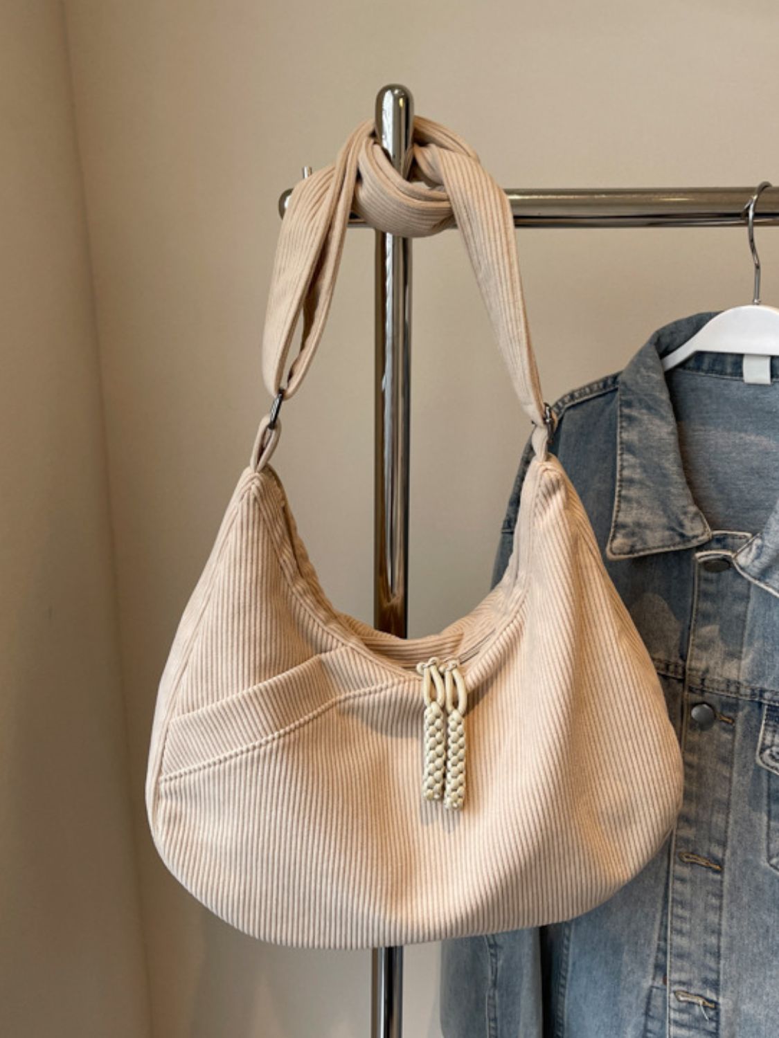 A large cream ribbed shoulder bag features a softly curved, slouchy silhouette, adjustable strap, and decorative braided pulls, styled beside a denim jacket.