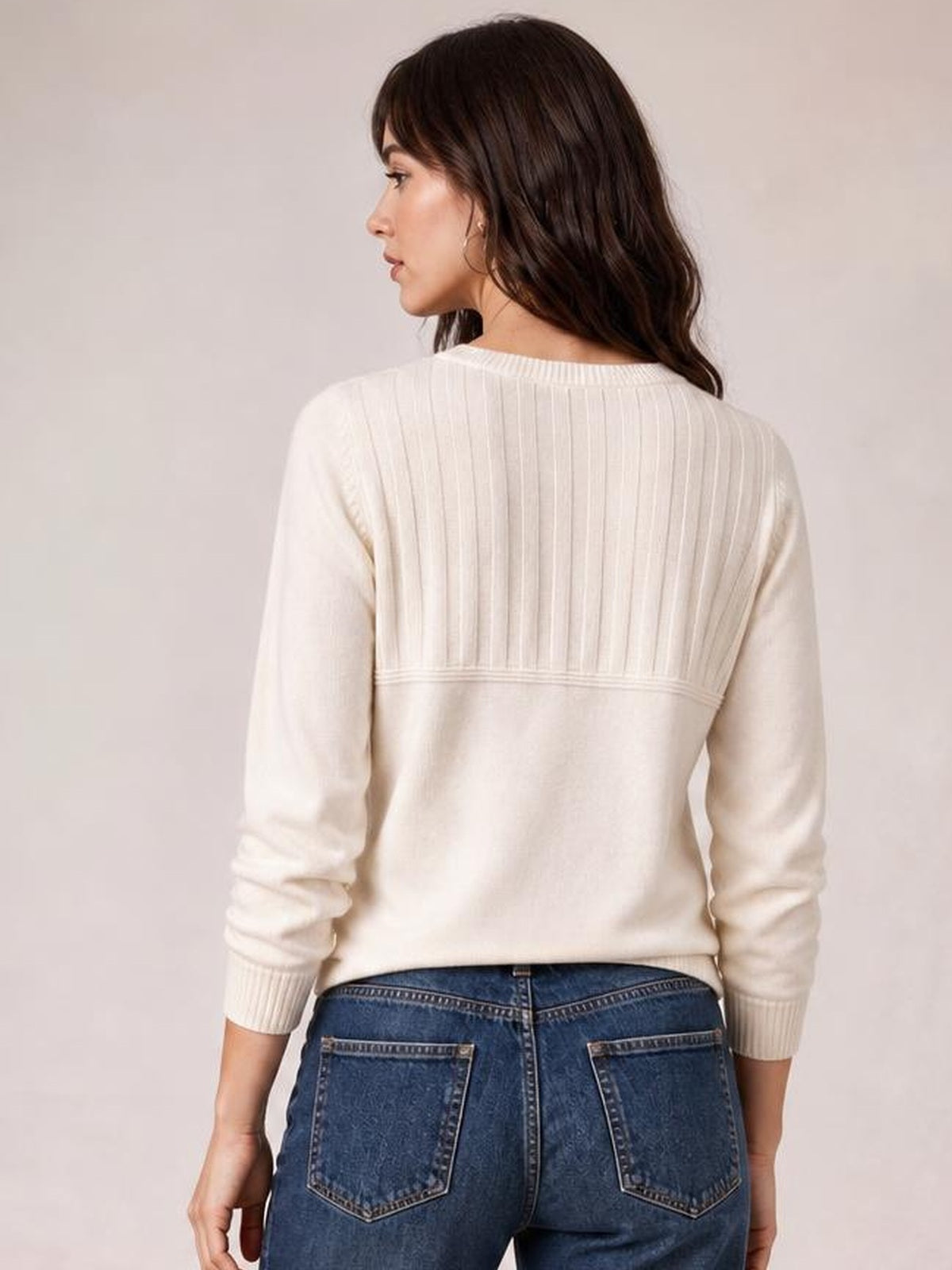 Model shown from the back in a cream ribbed sweater with a rounded neckline, long sleeves, vertical ribbed yoke, and horizontal seam detail, looking over her shoulder.