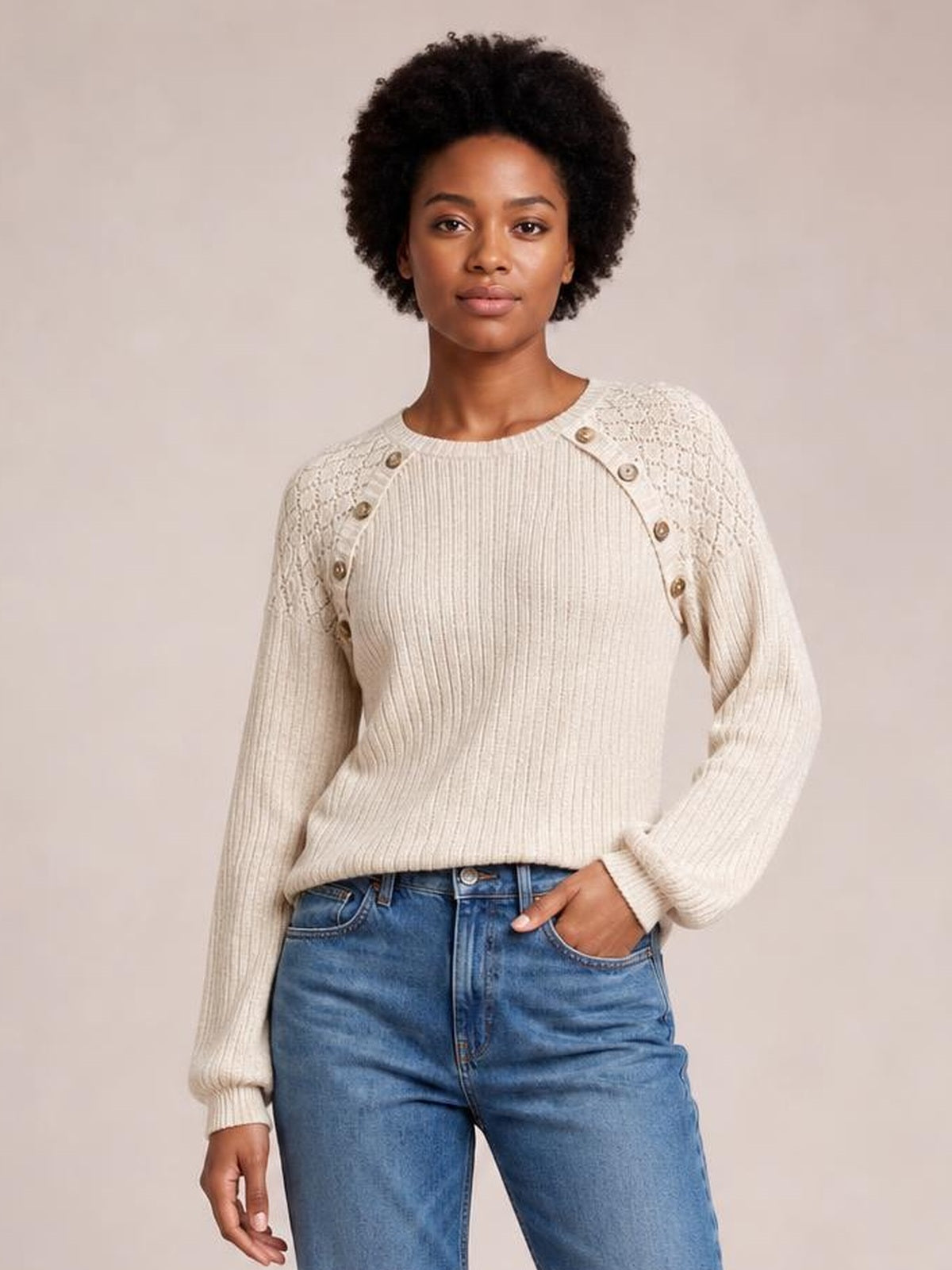A model poses with one hand in her jeans pocket wearing a cream ribbed round-neck top with decorative buttons and textured shoulder panels.
