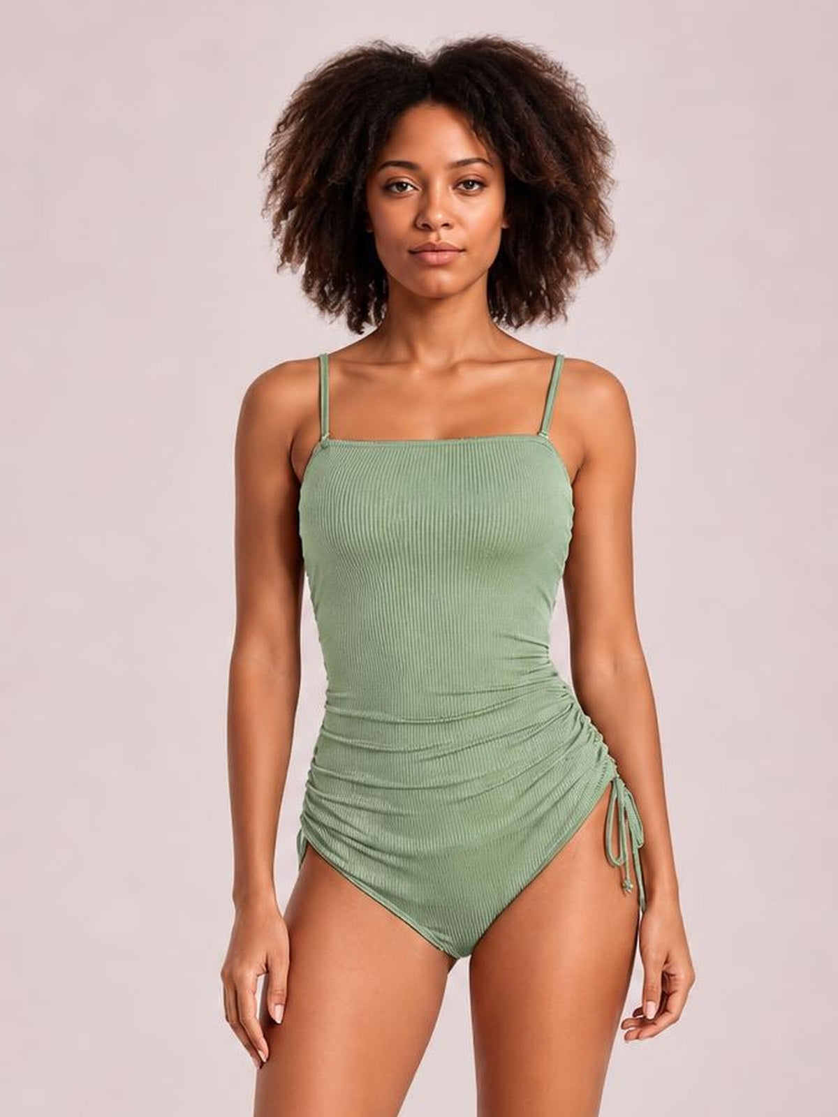 Model stands facing forward in a sage green ribbed one-piece swimsuit with thin straps, a square neckline, ruched sides, and a drawstring hip cutout.