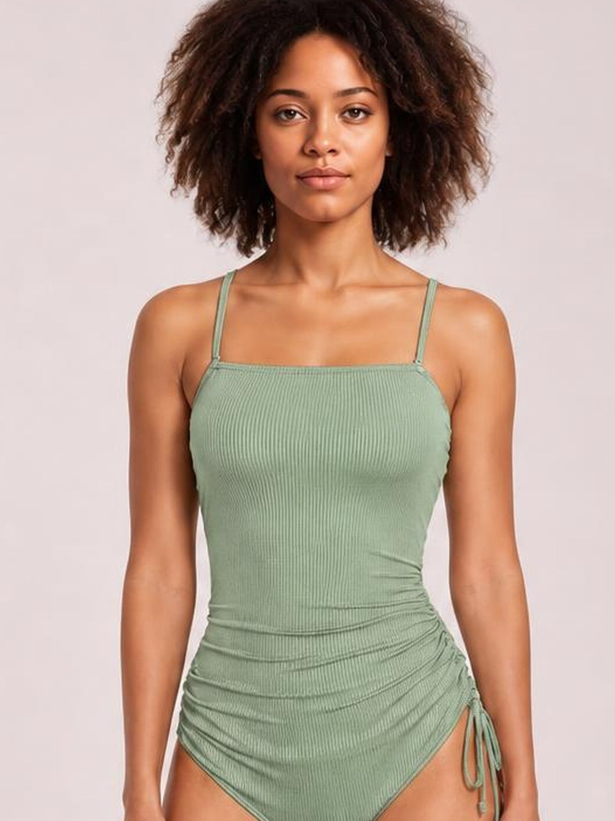 Model poses front-facing in a sage green ribbed one-piece swimsuit with thin straps, a square neckline, ruched drawstring sides, and cutout hips.