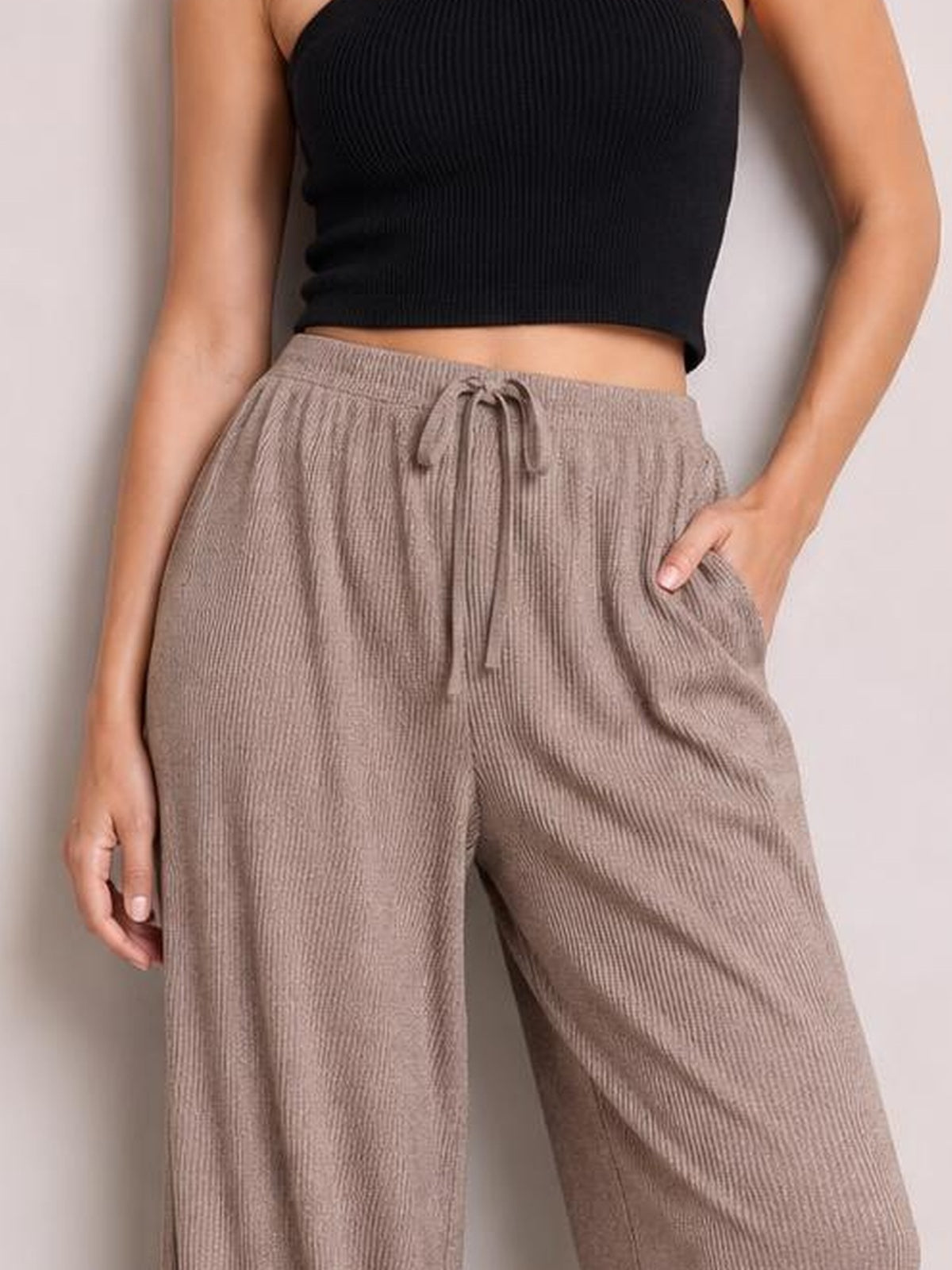 A model wears taupe ribbed drawstring wide leg pants with an elastic waistband and side pockets, posing with one hand tucked into a pocket.