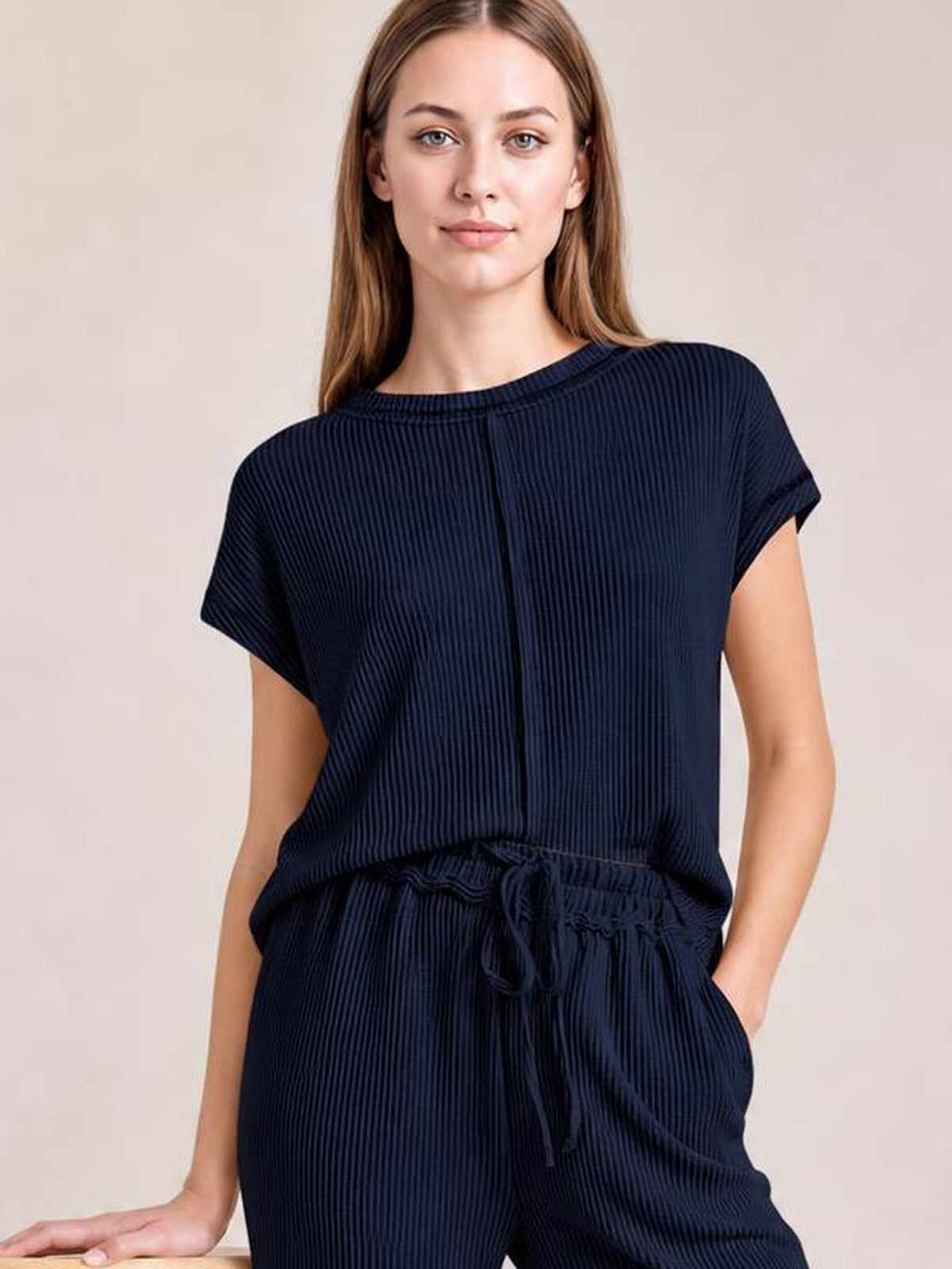 A model wears a navy ribbed top and pants set with short sleeves, exposed center seam, relaxed silhouette, drawstring waist, and side pockets.