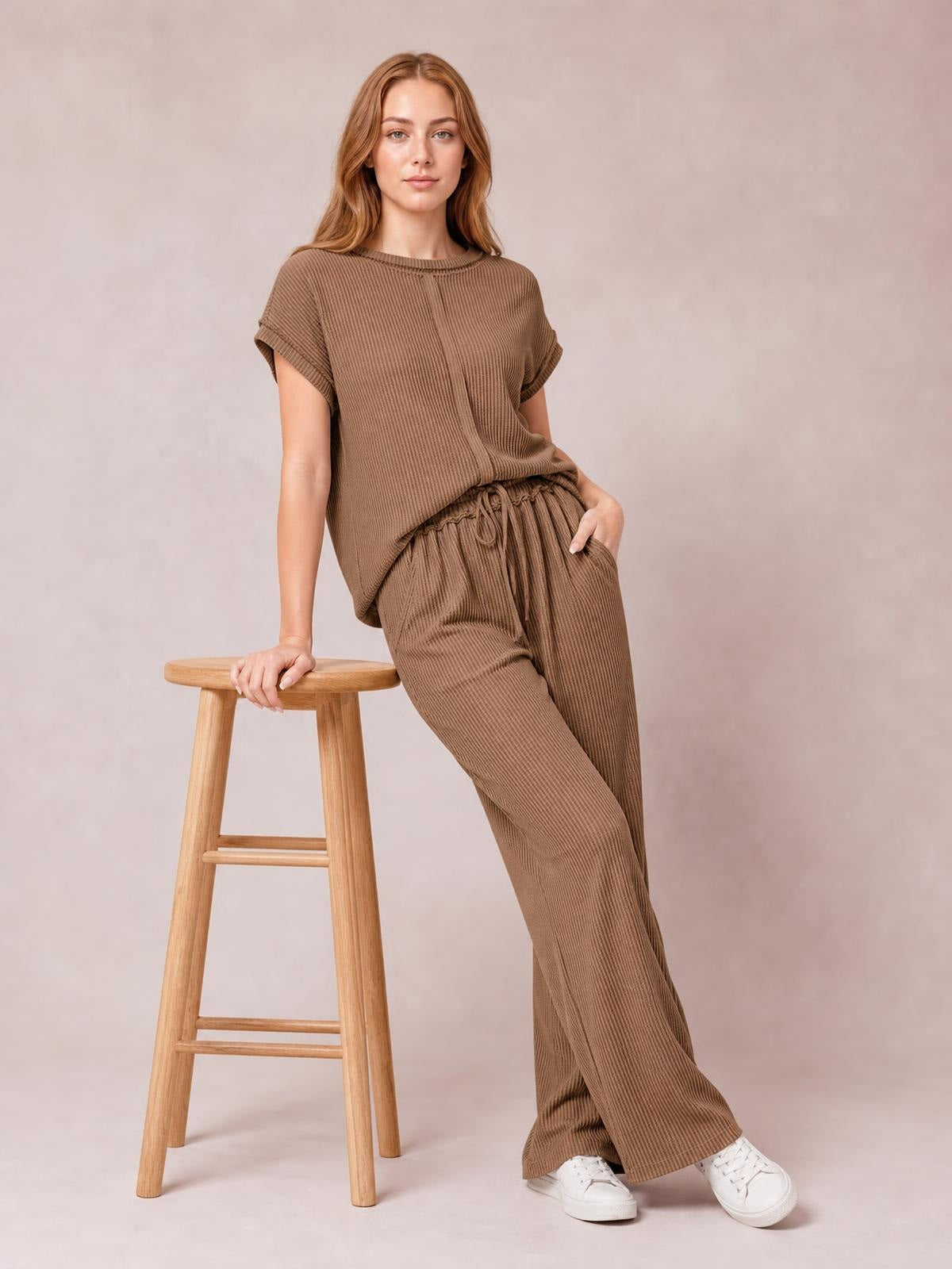 Model wearing a brown ribbed exposed seam top and wide-leg pants set, leaning against a stool in a relaxed pose with one hand in a pocket.