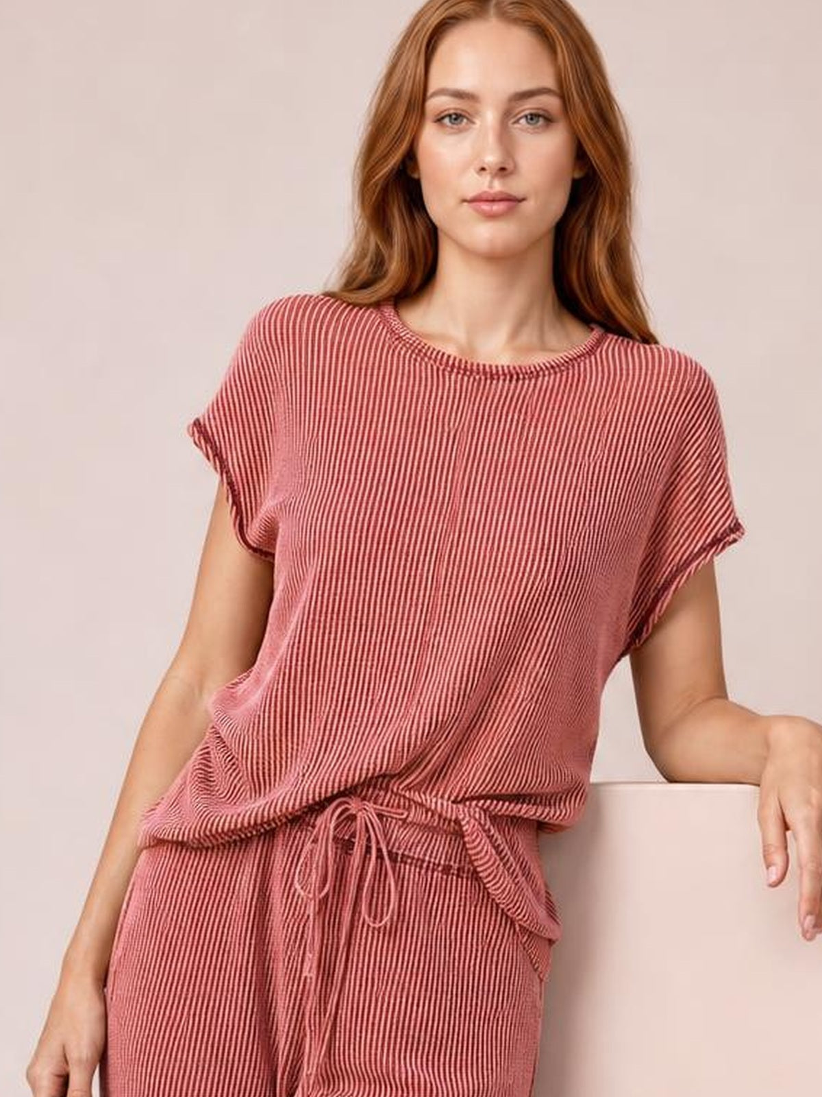 A model wears a dusty rose ribbed top and matching drawstring pants set, featuring short sleeves, exposed seam edges, and a relaxed silhouette.