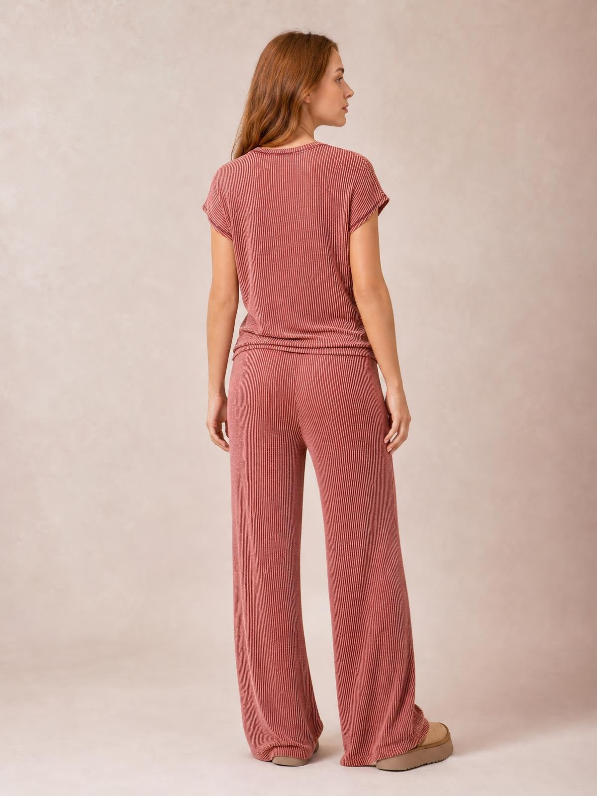 A model stands in profile from the back wearing a dusty rose ribbed top with exposed seams and relaxed wide-leg pants in a coordinated set.