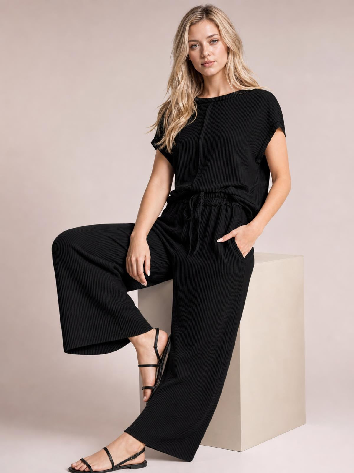 A model sits in a black ribbed exposed-seam top and matching wide-leg pants with a drawstring waist, posing with one leg raised.