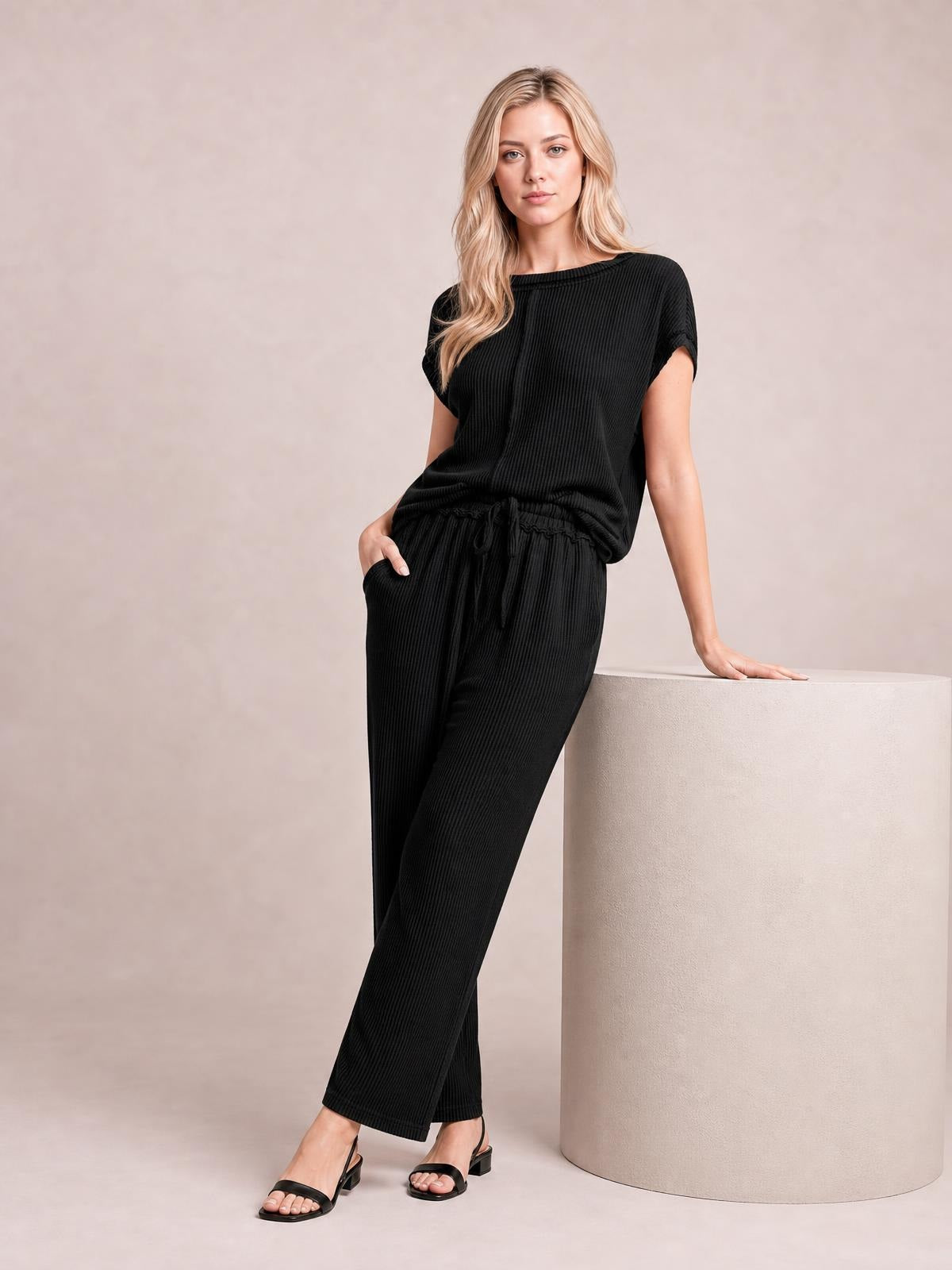 Model poses in a black ribbed exposed seam top and wide-leg pants set with a relaxed silhouette, elastic drawstring waist, and one hand in a pocket.
