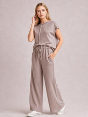A model poses in a taupe ribbed top and pants set with exposed seams, short sleeves, a drawstring waist, and relaxed wide-leg trousers.