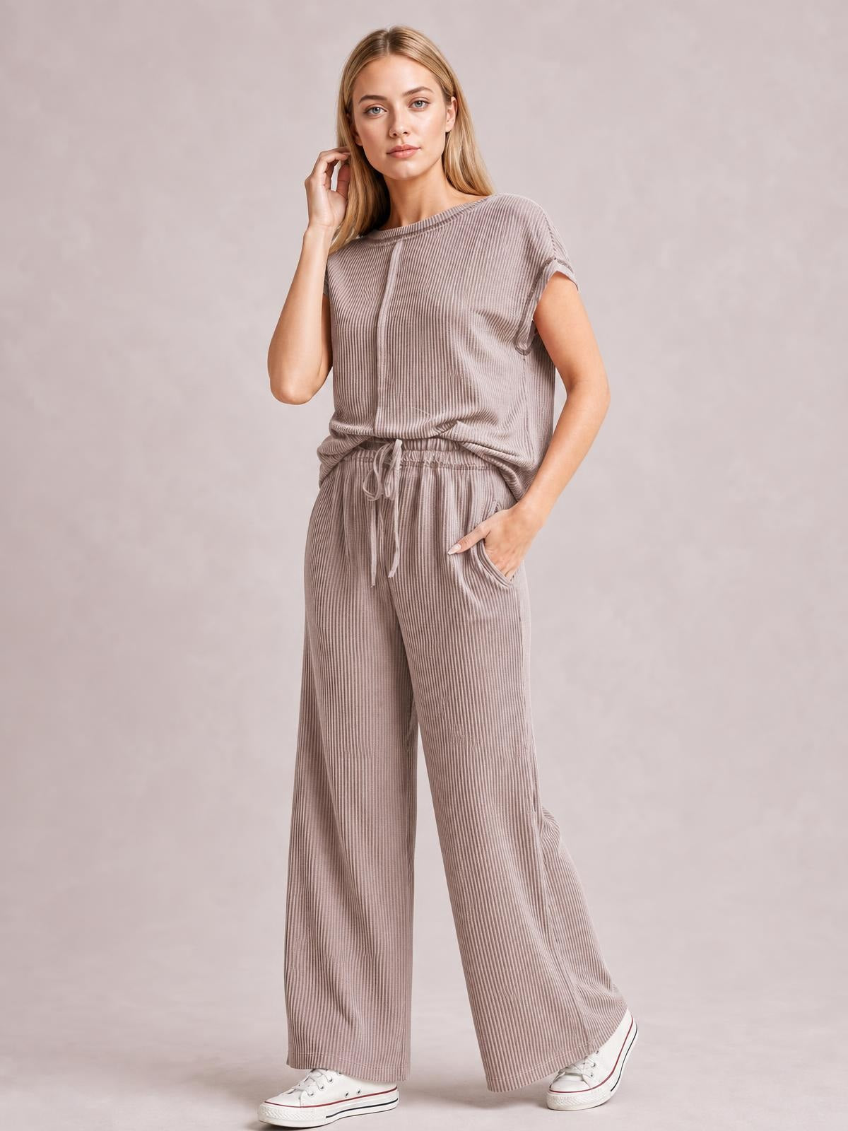 A model poses in a taupe ribbed top and pants set with exposed seams, short sleeves, a drawstring waist, and relaxed wide-leg trousers.