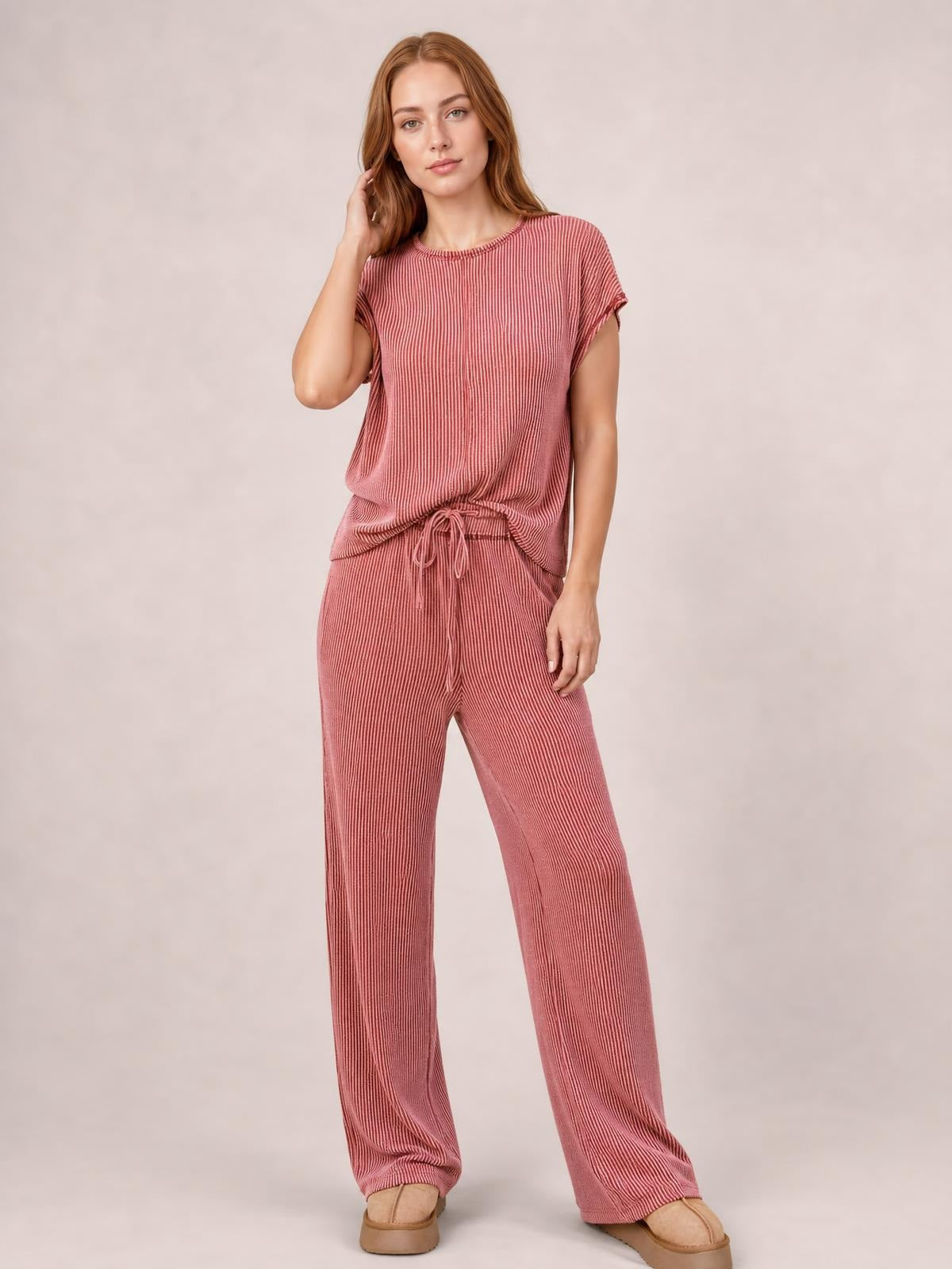 A model wears a dusty rose ribbed top and wide-leg pants set with short sleeves, exposed seam detailing, and a drawstring waist, posing with one hand in her hair.