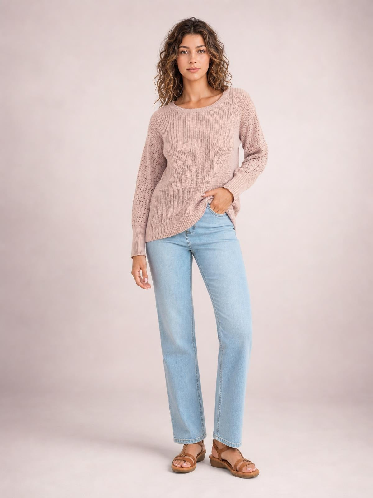 A model wears a blush pink ribbed round-neck pullover sweater with long openwork sleeves and dropped shoulders, posing with one hand in her jeans pocket.