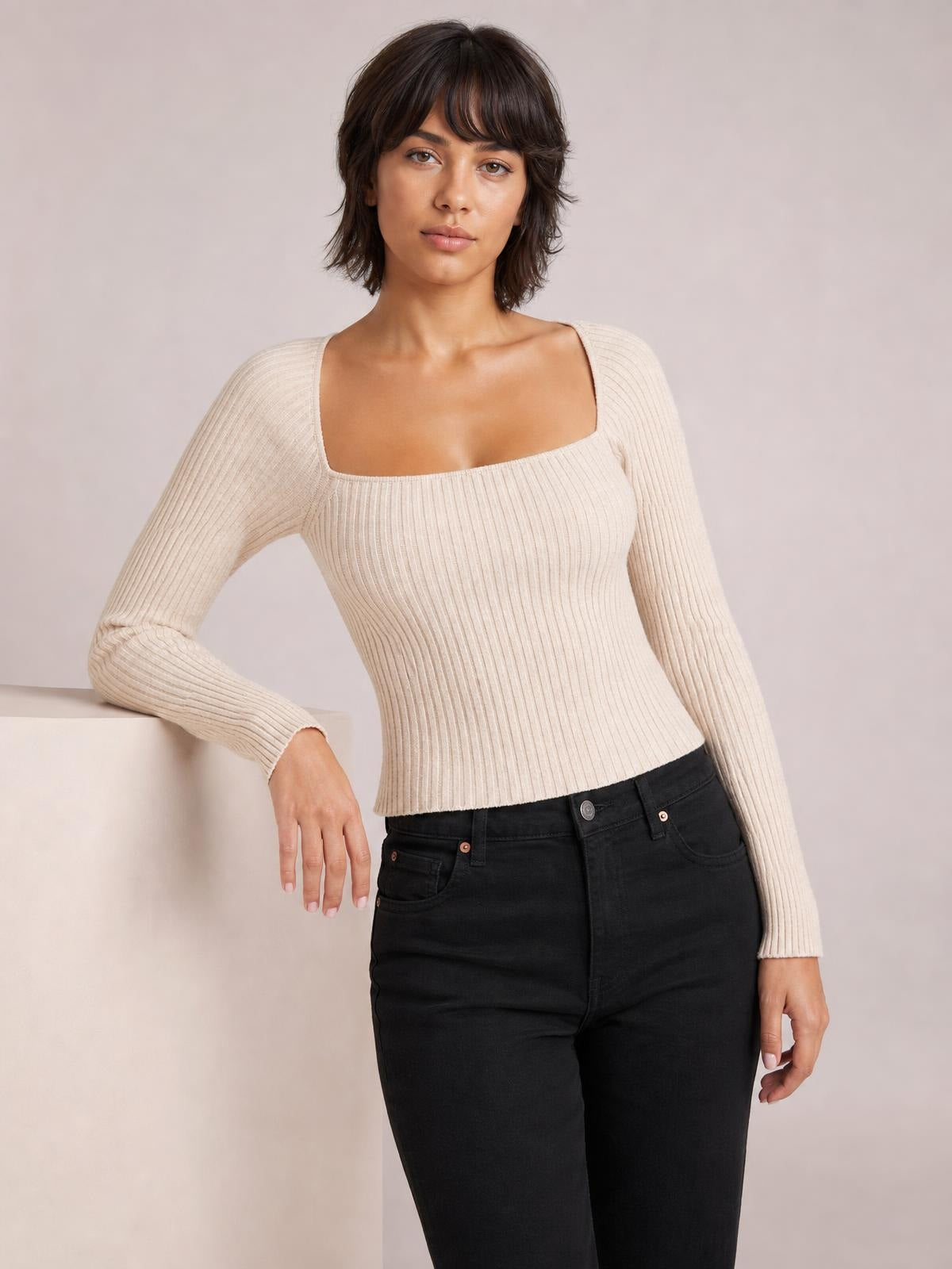 Model wearing a cream ribbed square neck long sleeve sweater with a fitted silhouette, stretch knit construction, and one arm resting on a ledge.