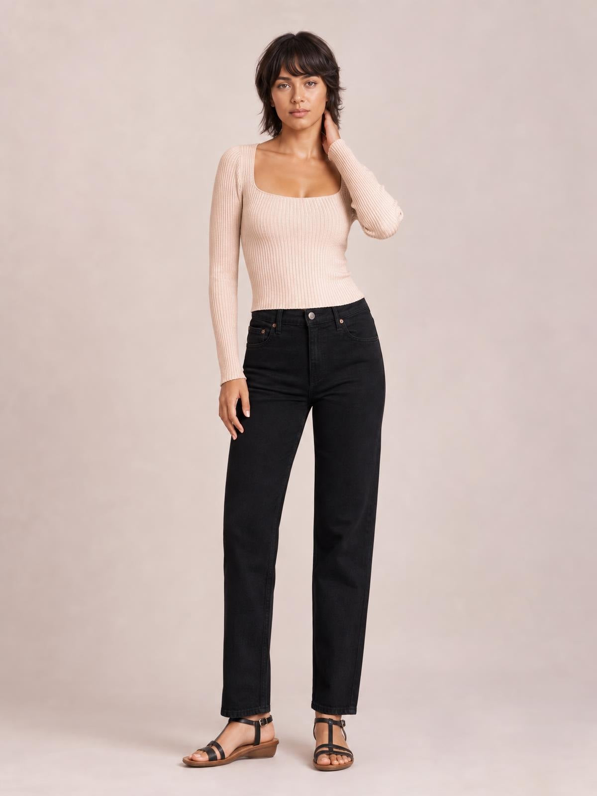 Model wearing a blush pink ribbed square neck long sleeve sweater with a fitted silhouette, standing with one hand touching her neck.